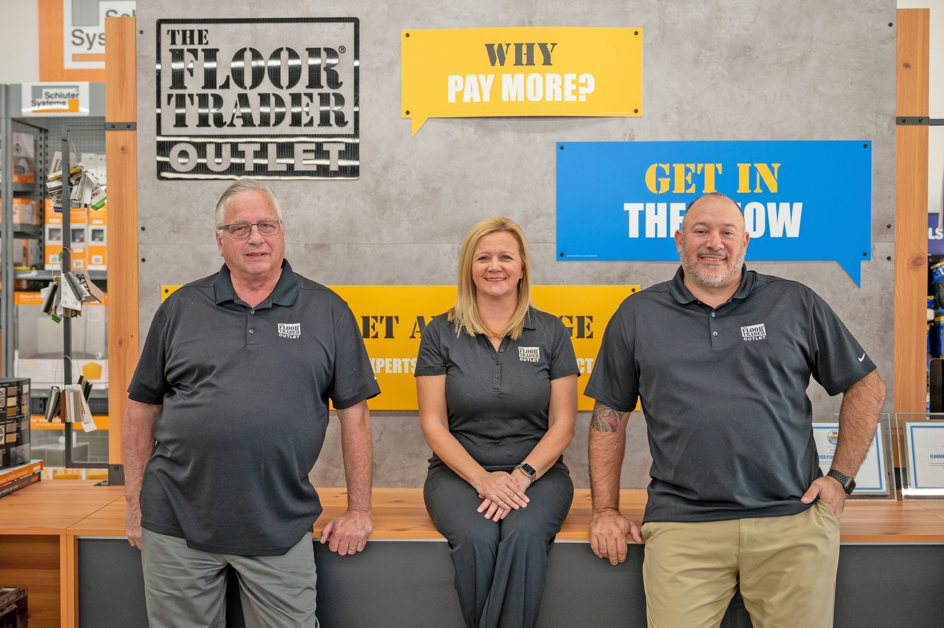 Three people in polo shirts stand behind a wooden counter at The Floor Trader Outlet in front of promotional signs.