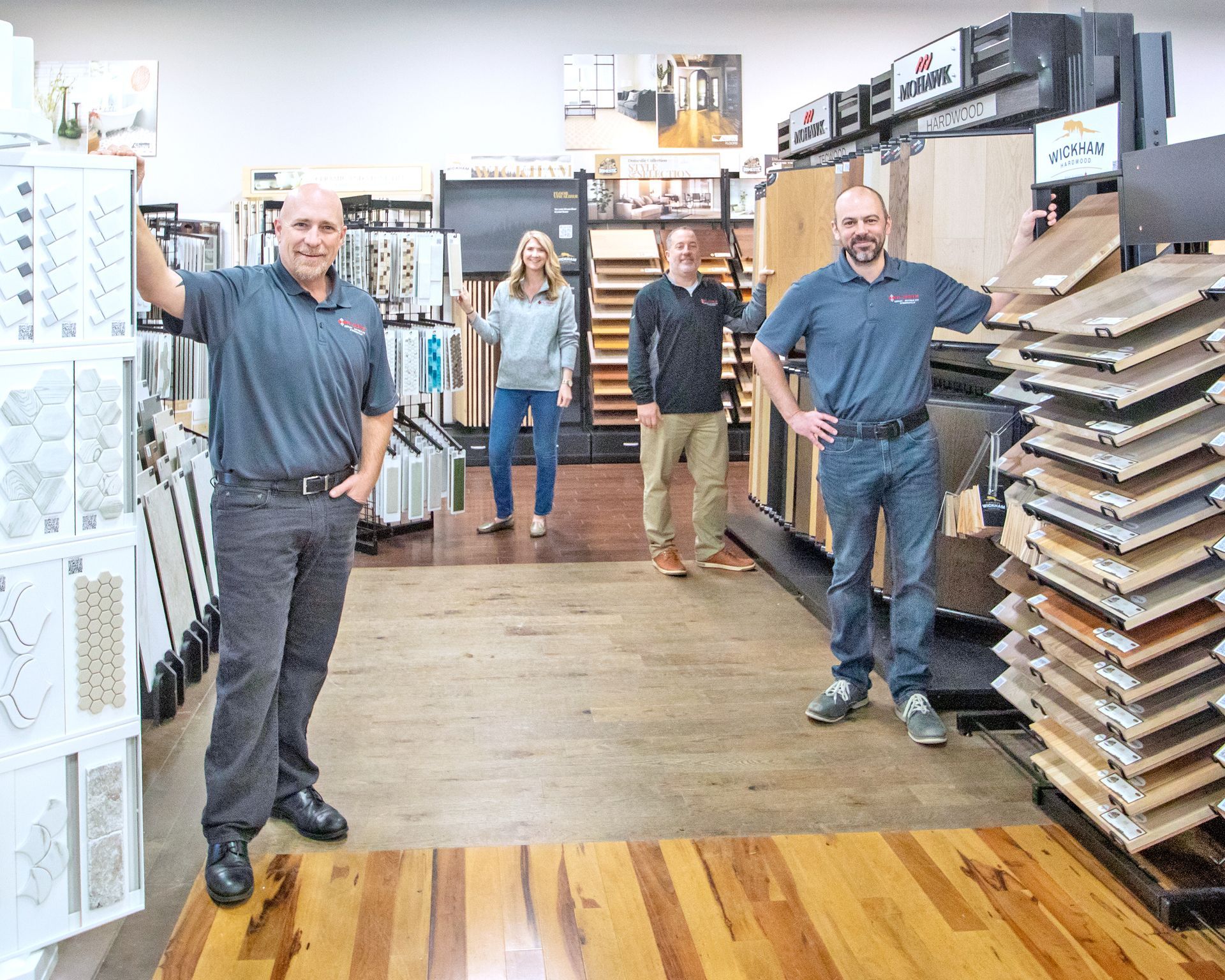Four people stand in a flooring showroom, posing near displays of tile and wood samples on shelves.