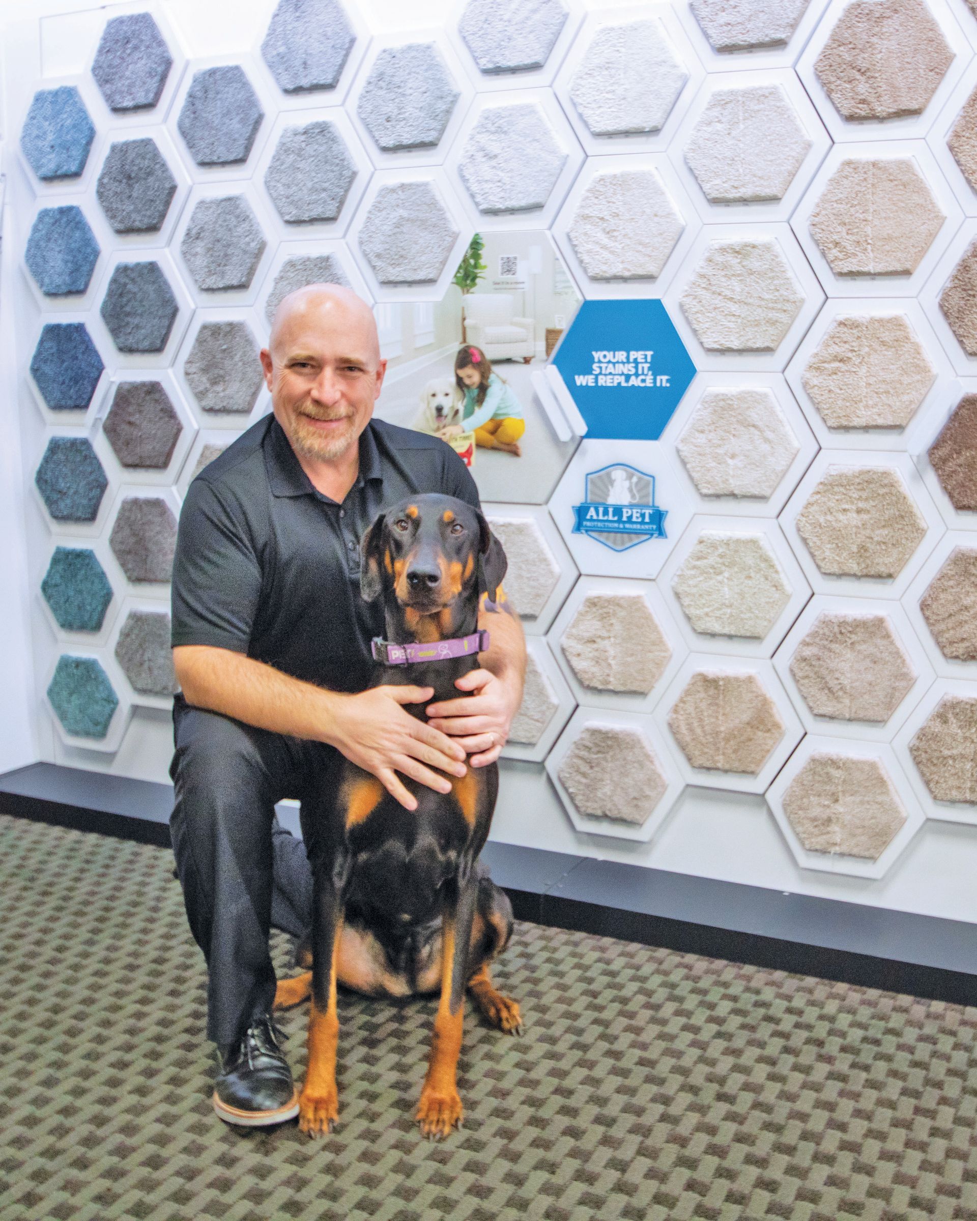 A smiling person kneels next to a black and tan dog in a store, with hexagonal carpet samples displayed on the wall behind.