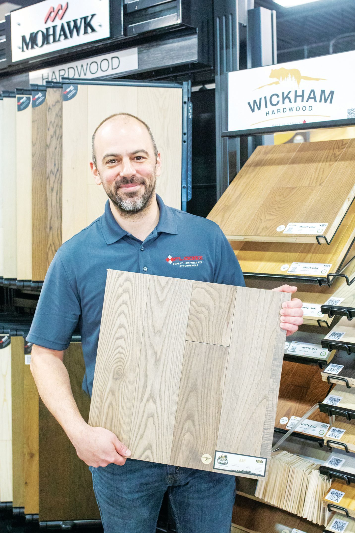 A person holding a light-toned wood flooring sample in a showroom with display racks of flooring options.