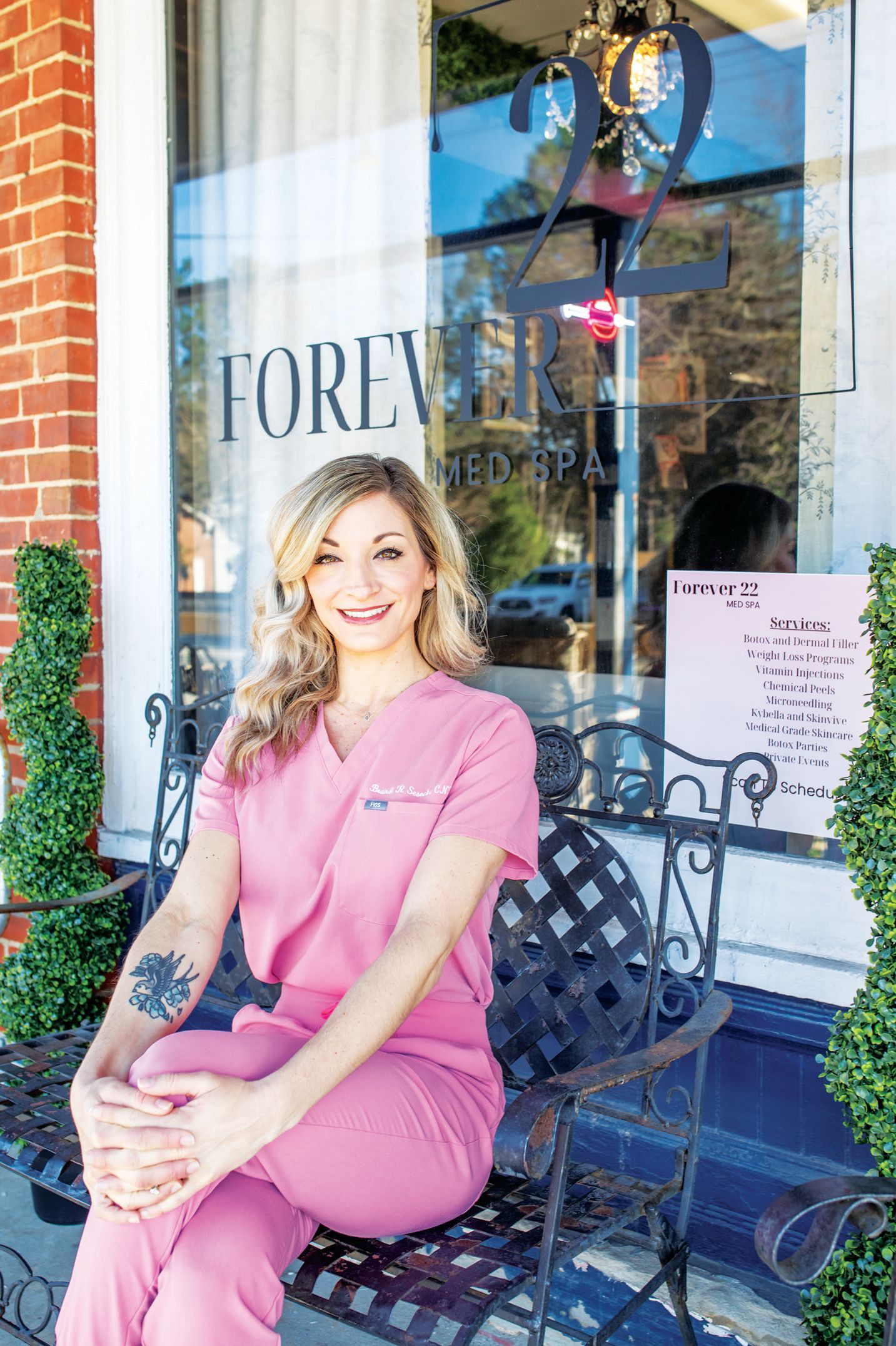 A person in pink scrubs smiling while sitting on an ornate black bench in front of a shop window labeled