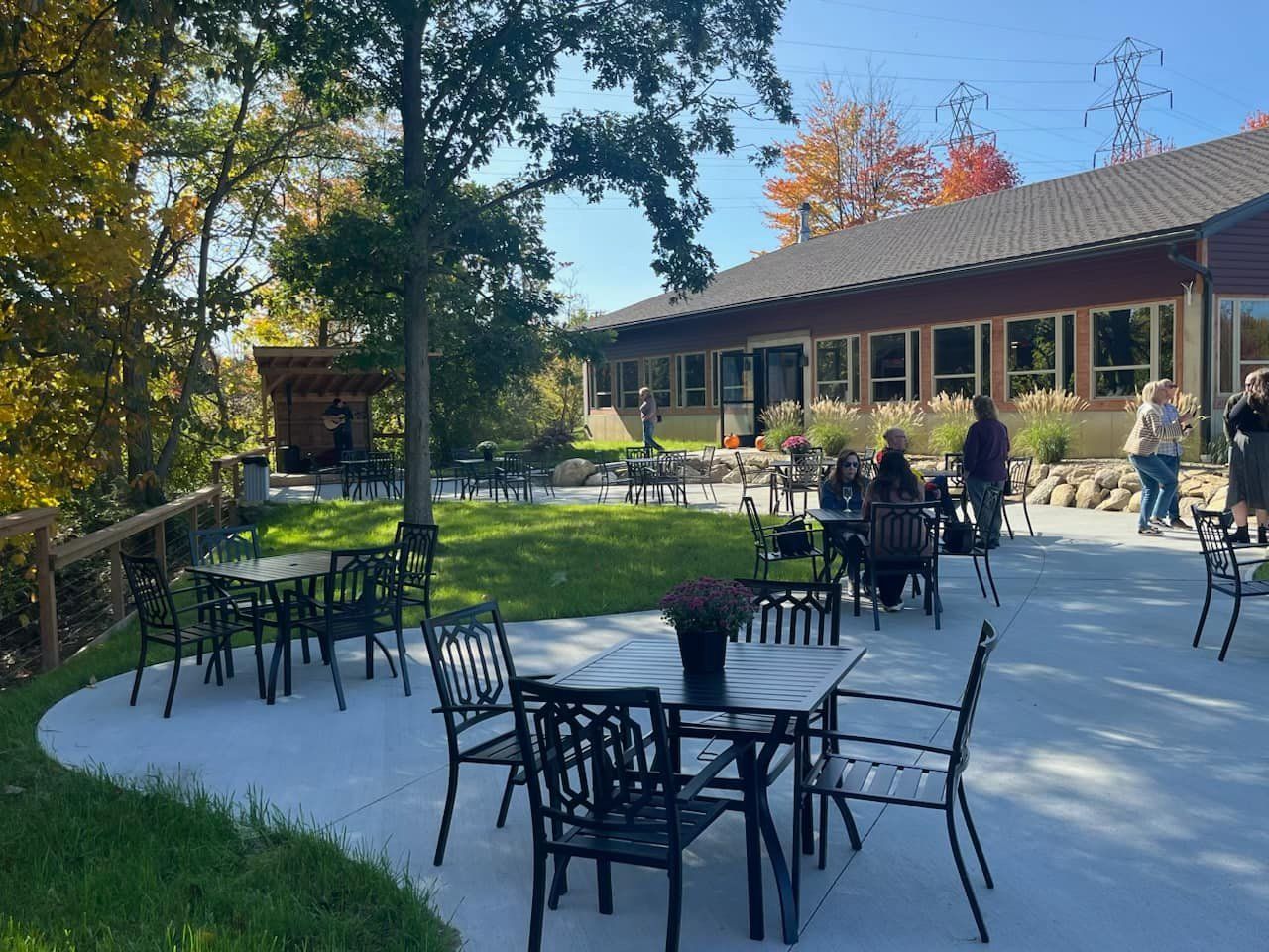 Patio area with black metal tables and chairs outside a building on a sunny day with trees in the background.