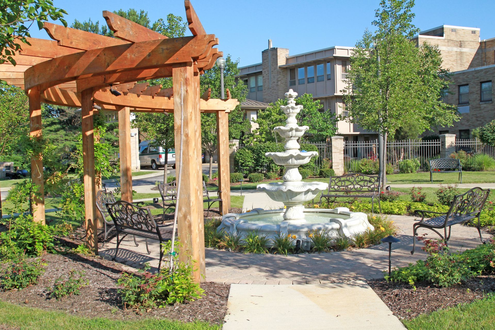 A tiered white fountain sits in a garden plaza with a wooden pergola, benches, and a building in the background.