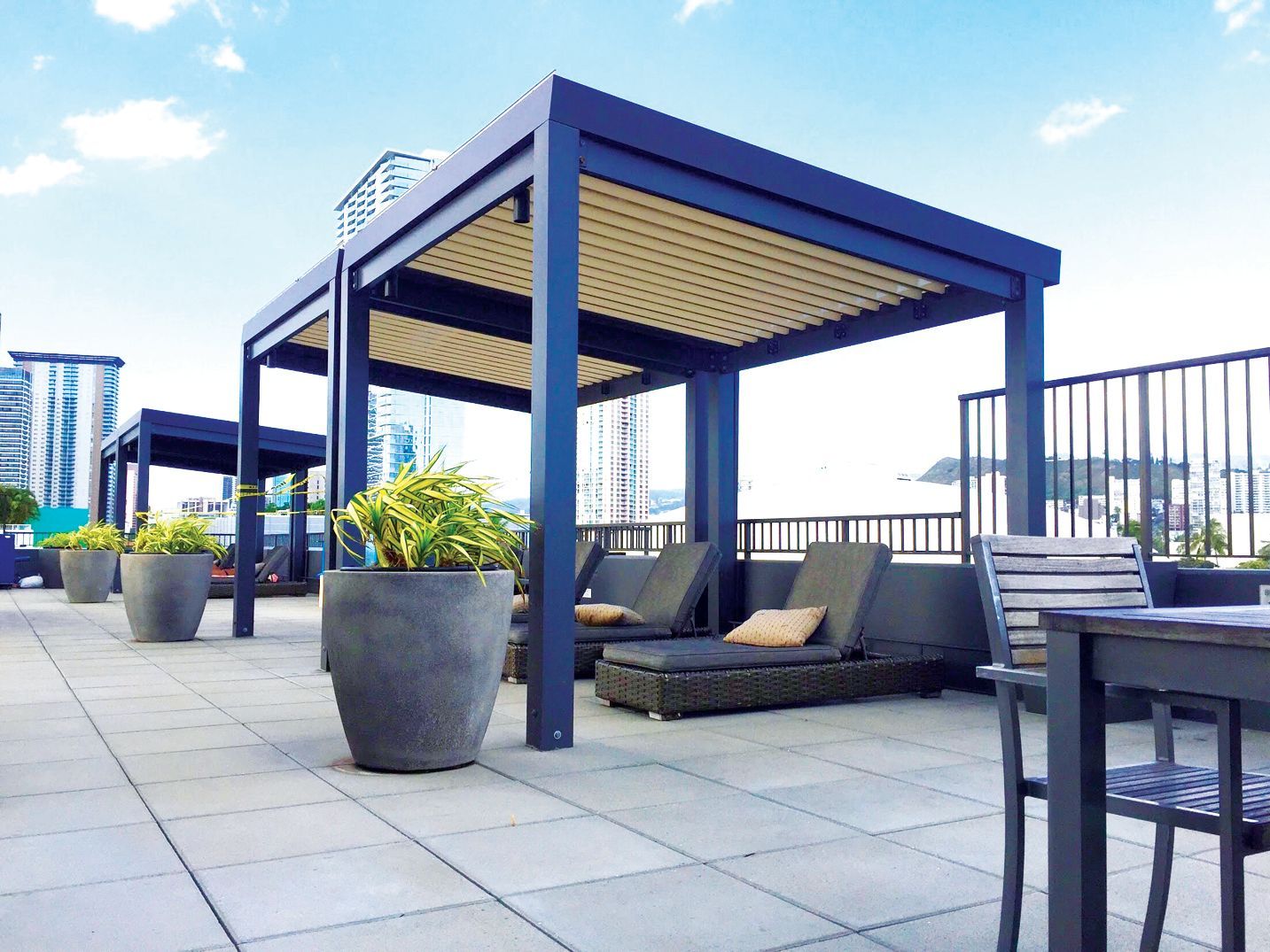 A rooftop lounge featuring dark metal pergolas with slatted roofs, potted plants, and lounge chairs on tiled flooring.