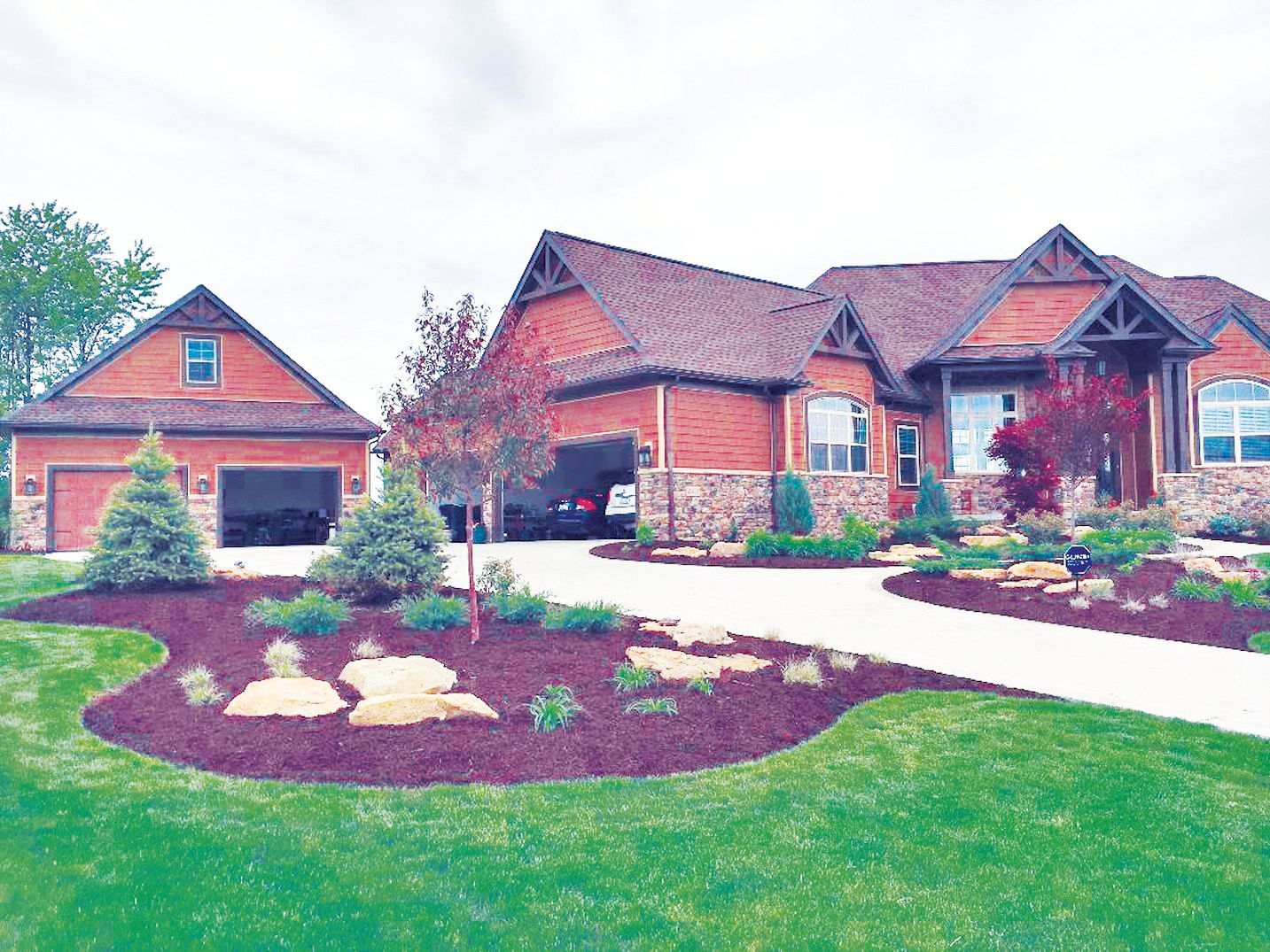 A brick house with a detached garage, a curved mulched garden bed with boulders, and a green lawn under a cloudy sky.