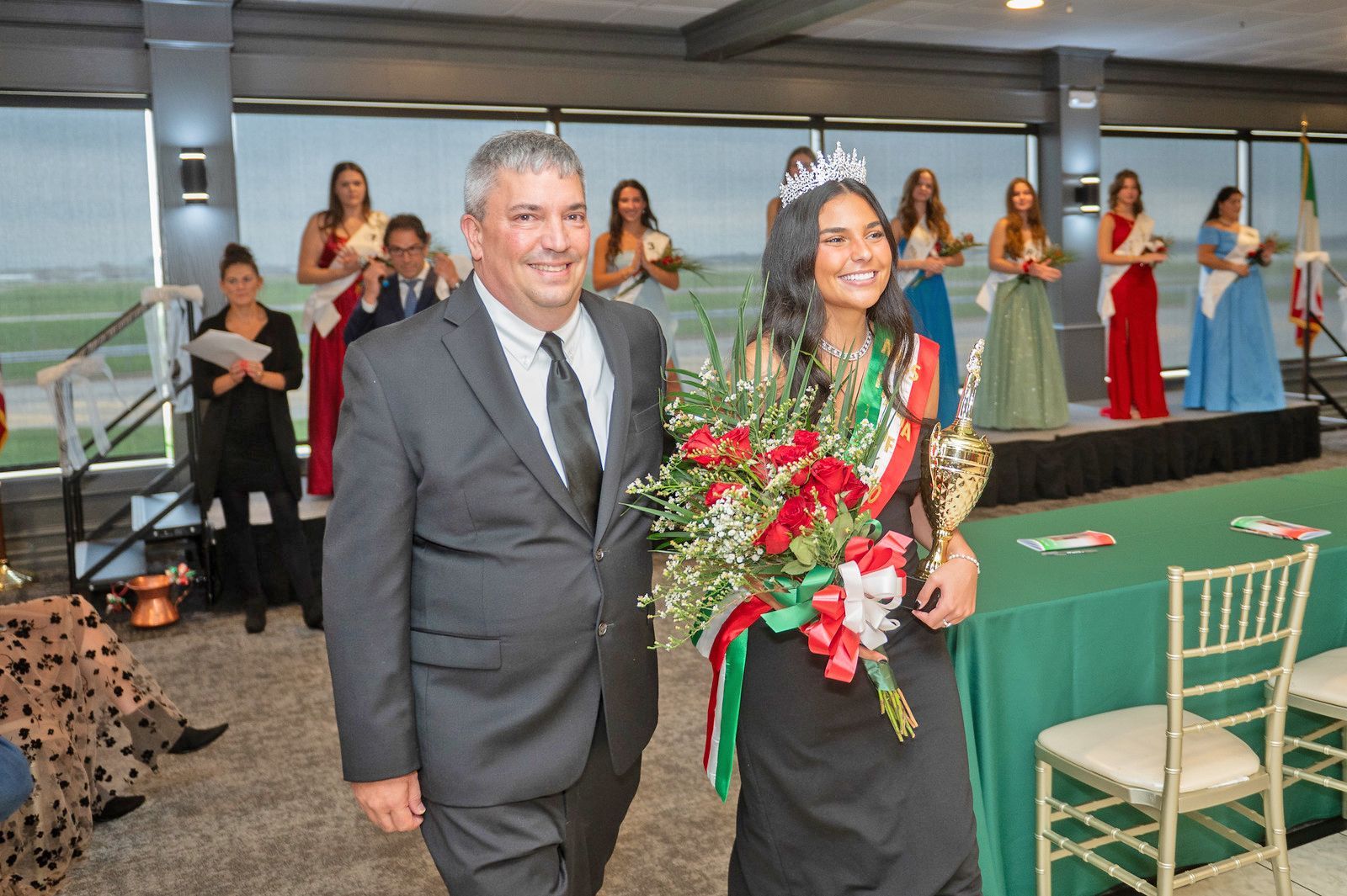 A smiling person in a crown and sash holds a bouquet and trophy, walking next to a person in a suit in a banquet room.