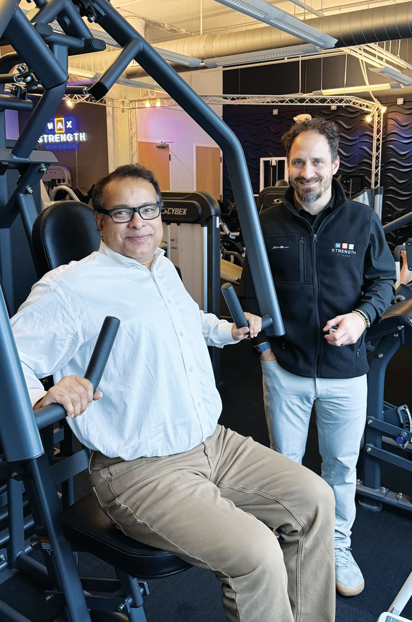 Two men in a gym, one seated on a chest press machine and the other standing beside him, both smiling.