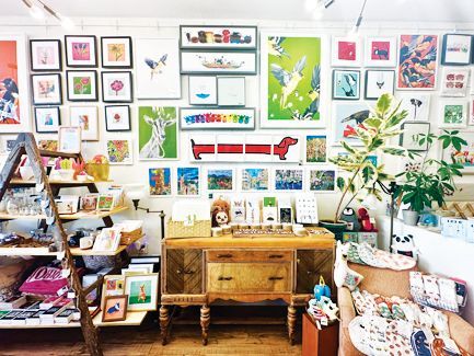 A shop wall covered in framed colorful art prints, with a wooden sideboard, ladder shelving, and plants in the foreground.