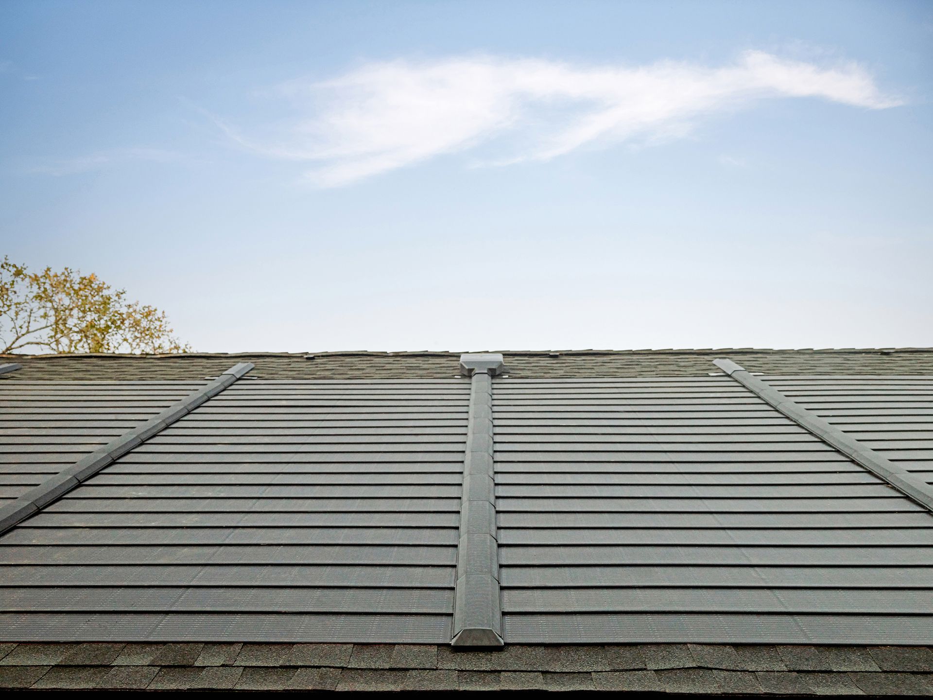 A rooftop view of dark gray solar roof shingles with metal vertical channels against a blue sky with light clouds.