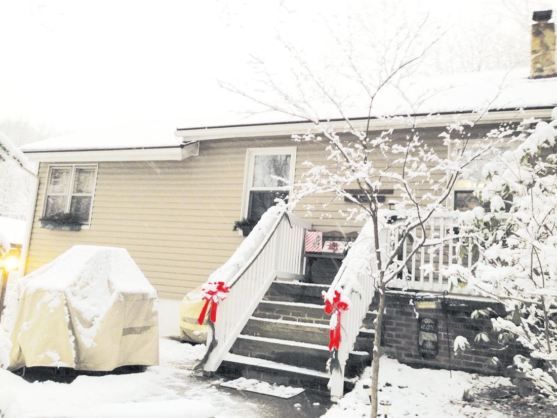 A snowy backyard scene featuring a tan house with a small staircase adorned with red bows and a grill covered in a tarp.