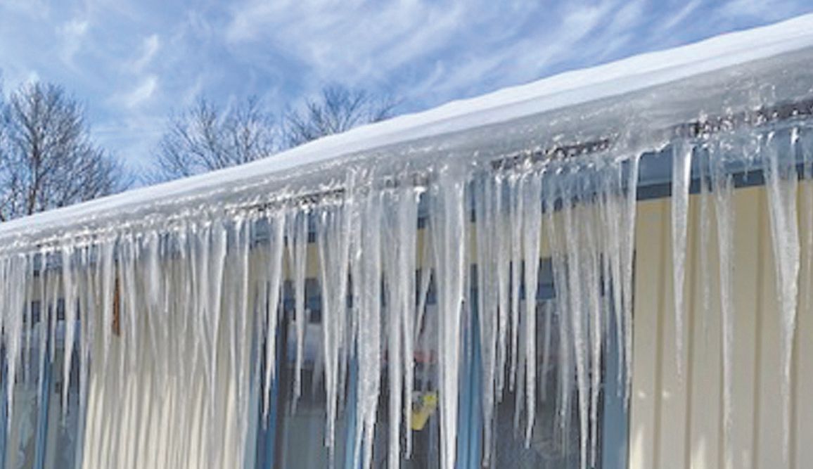 Long icicles hanging from the roofline of a building with snow on the roof against a blue sky with wispy clouds.