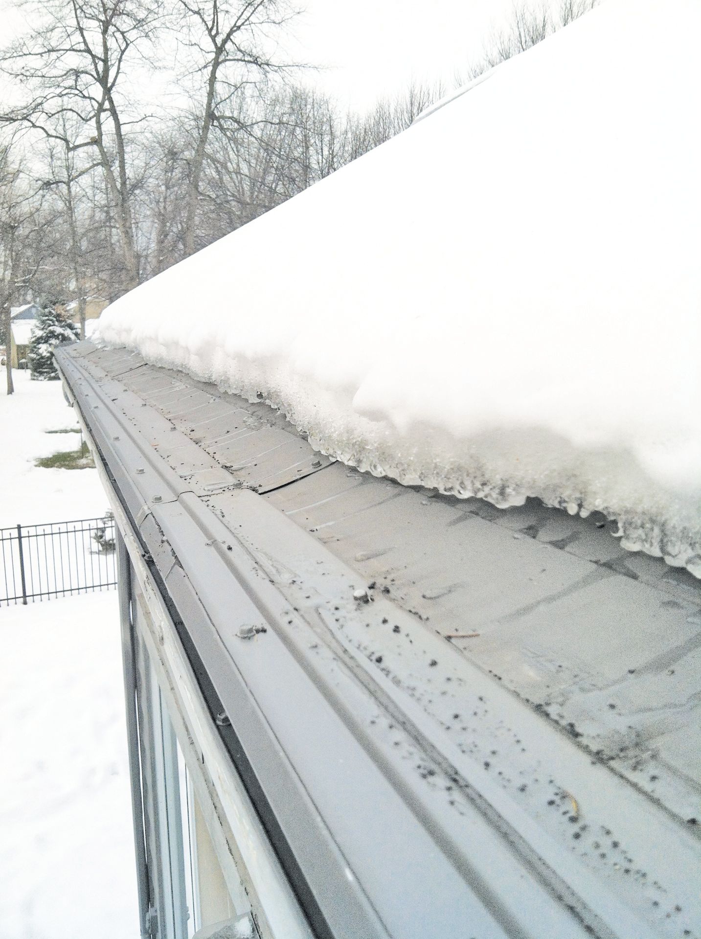 A close-up view of a metal gutter cover partially covered by a thick layer of snow and icicles on a residential roof.