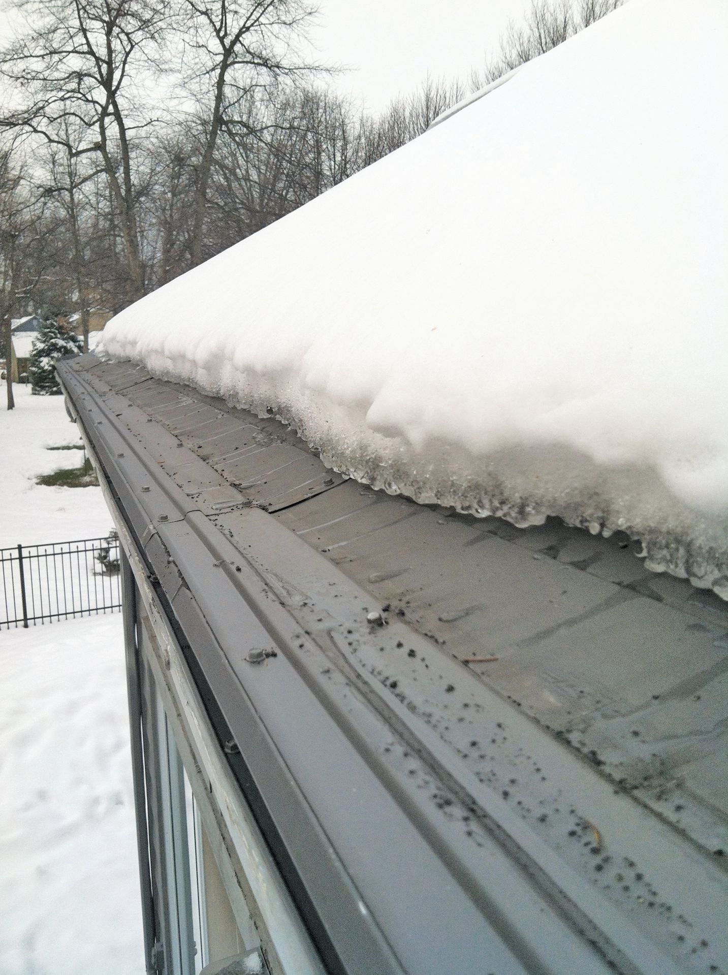 A thick layer of snow sits on a grey roof, with icicles hanging from the edge above a gutter.