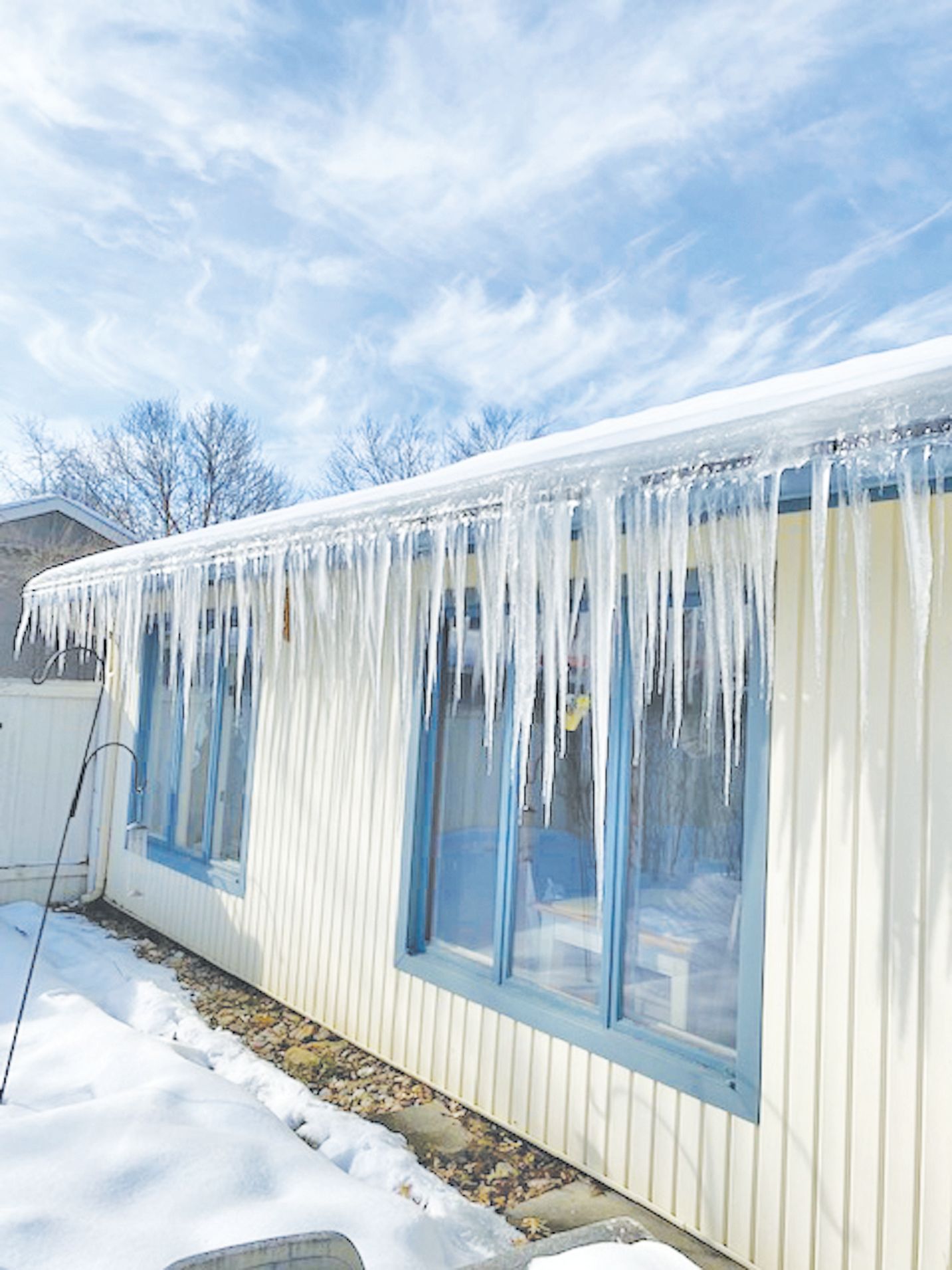 Long, sharp icicles hang from the snowy eaves of a pale yellow house with blue-trimmed windows on a sunny winter day.