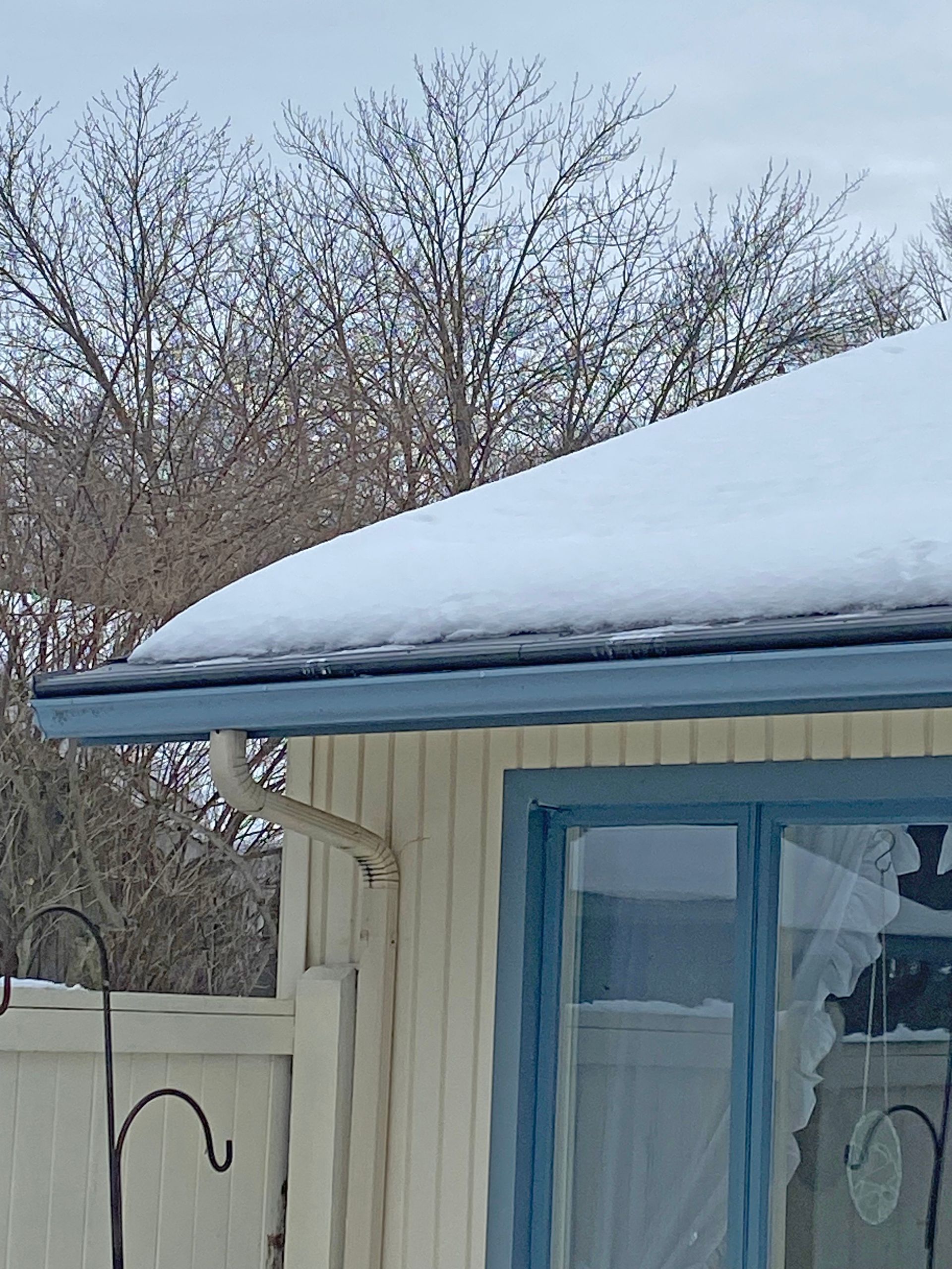 A snow-covered roof with blue gutters and trim above a light-colored wood-paneled wall and window, with trees in the back.