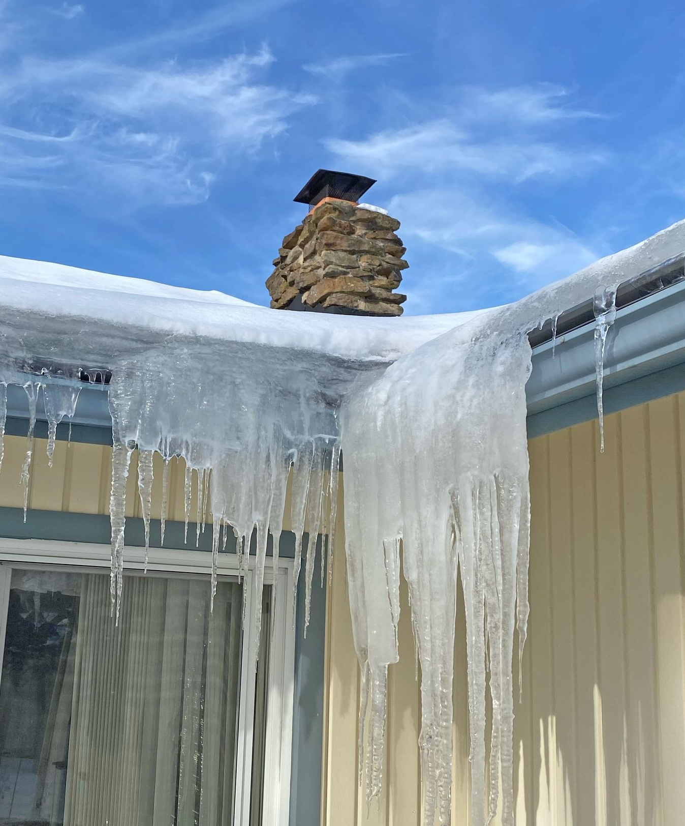 Large icicles hang from the snowy roof edge of a yellow house with a stone chimney under a bright blue sky.