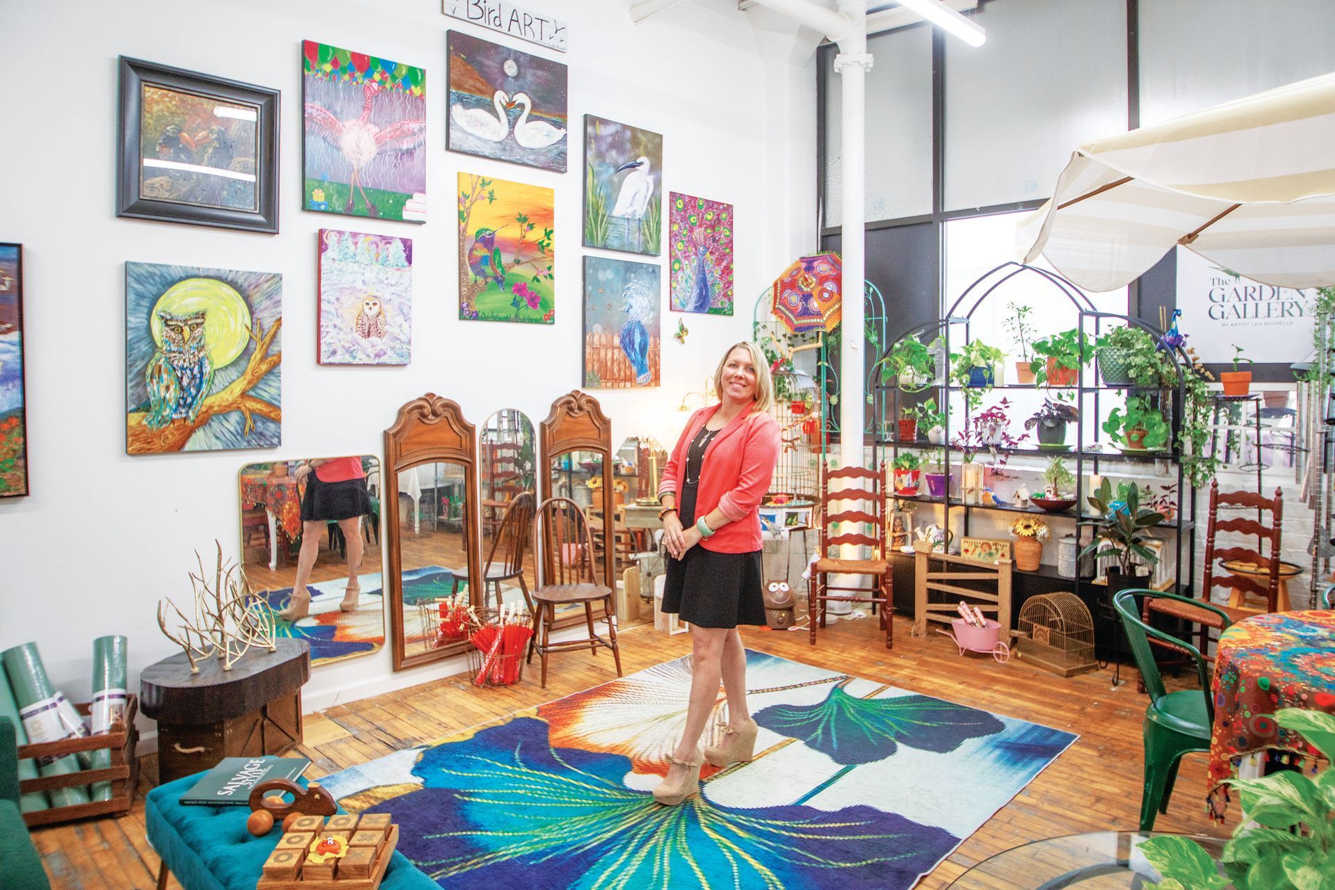 A person stands in a brightly lit art gallery, surrounded by colorful paintings on white walls and indoor plants.