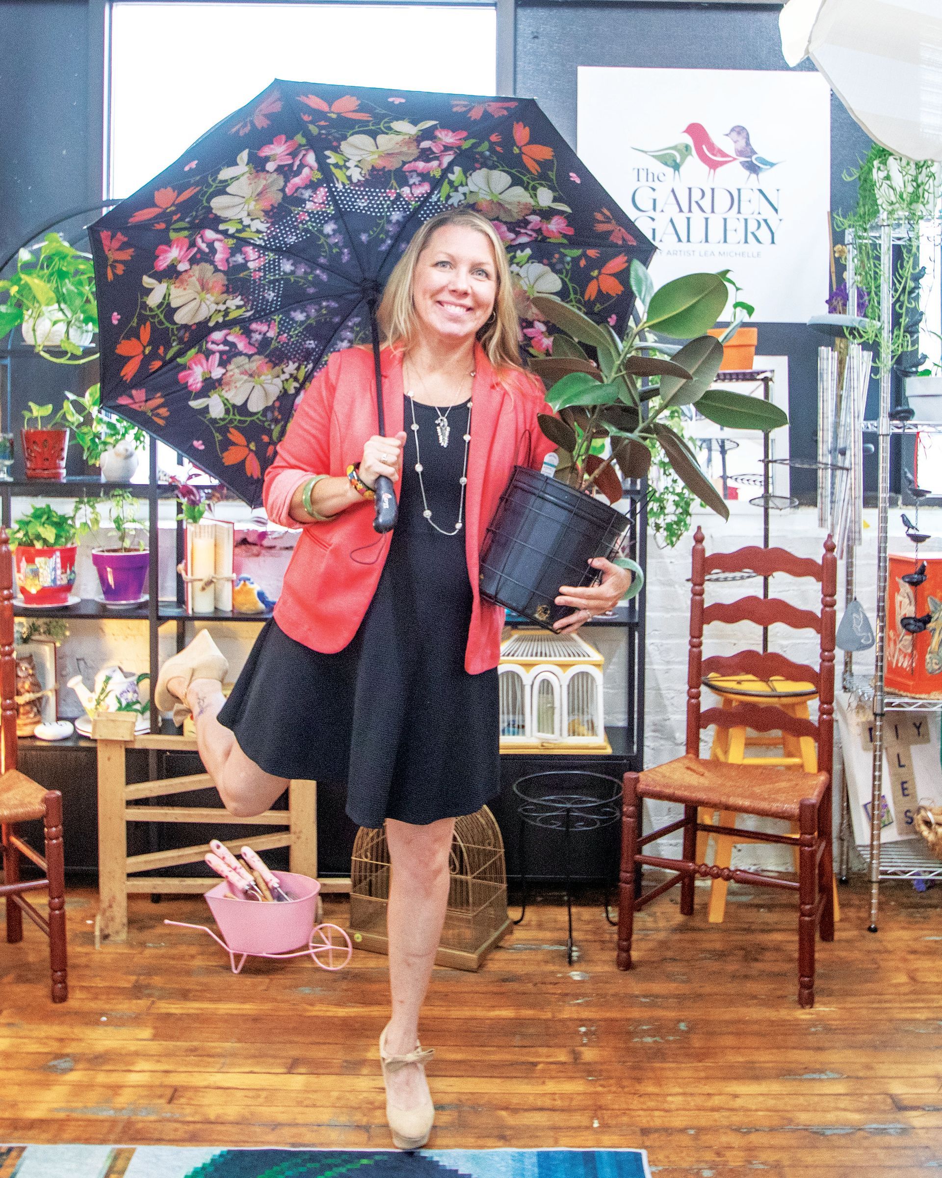 Smiling person in a coral blazer holding an umbrella and a potted plant in a garden decor shop.