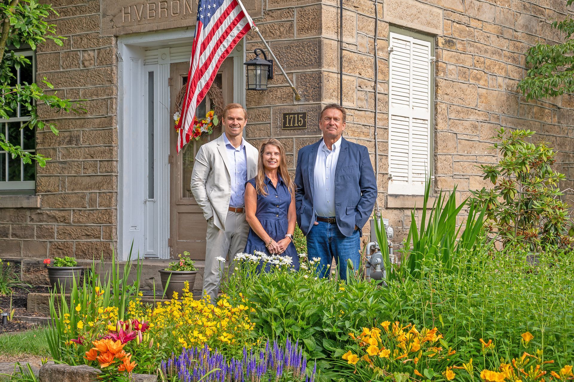 Three people stand in front of a stone building with an American flag above the entrance and a garden in the foreground.