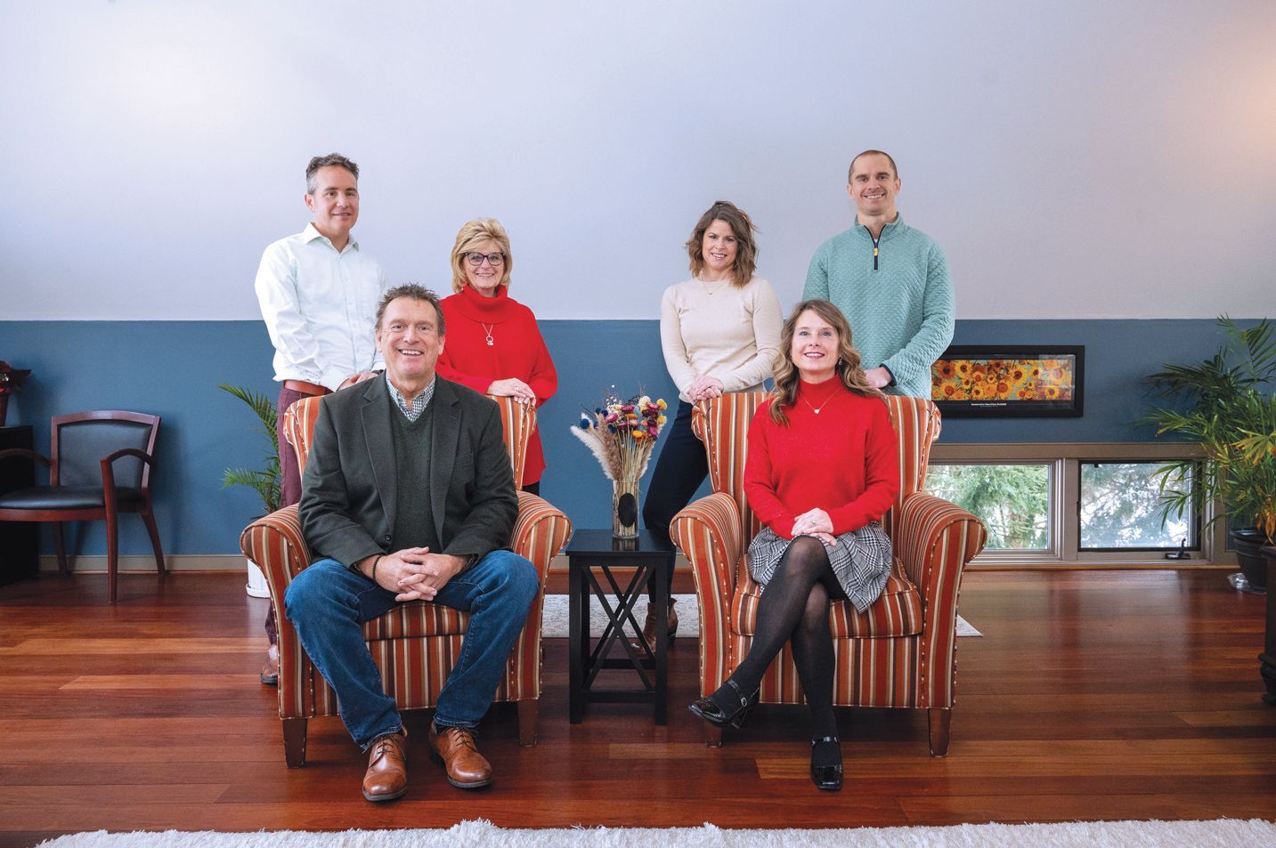 Six people stand or sit in an indoor room with blue walls, wood floors, and a fireplace, posing for a group portrait.