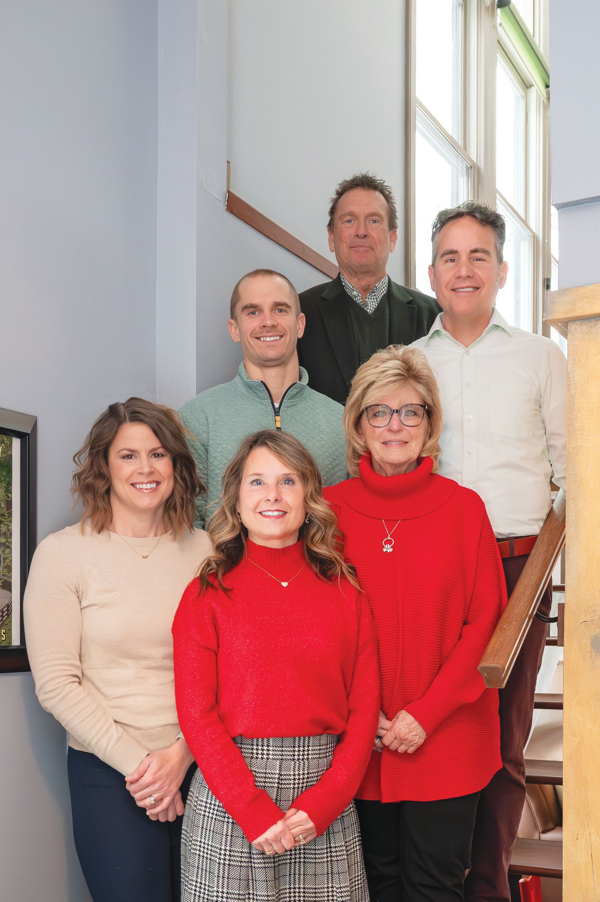 Six people pose for a portrait on a staircase, wearing casual attire, with neutral expressions in a brightly lit room.