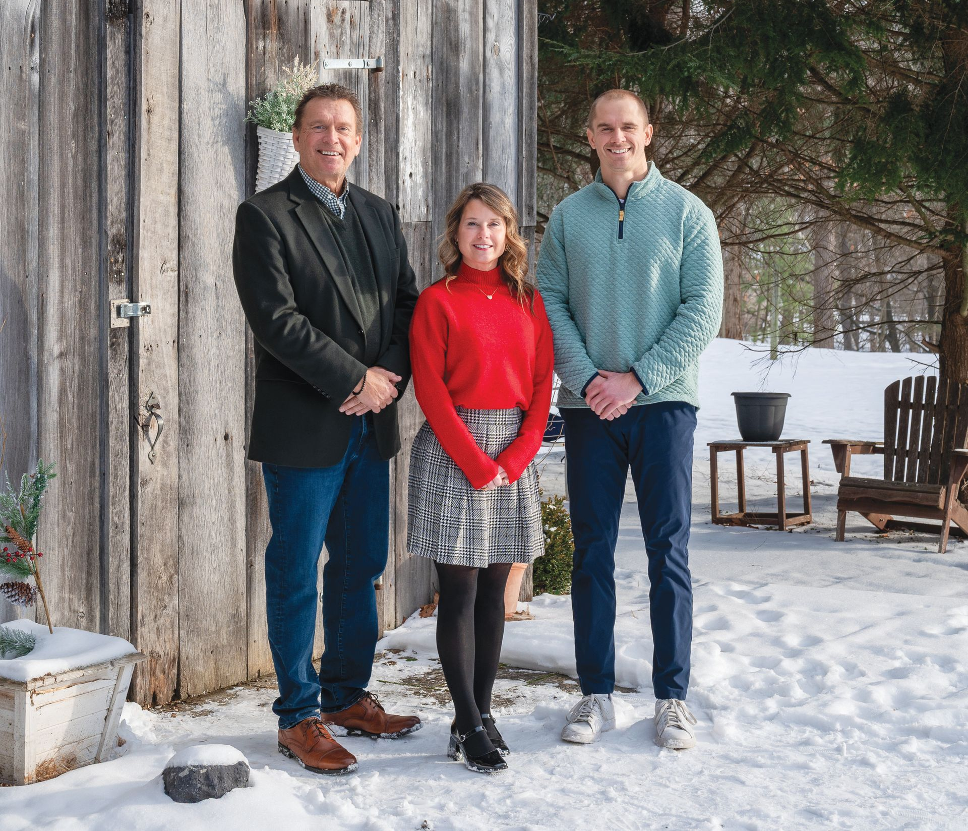 Three people pose together outside in the snow in front of a rustic wooden structure, smiling at the camera.