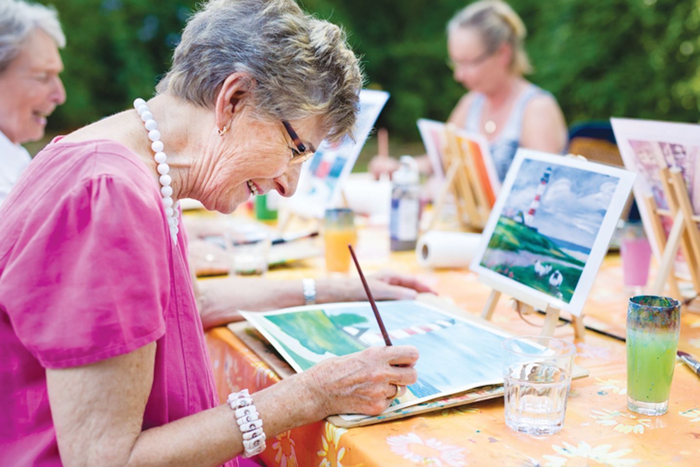 People sit at a table outdoors, happily painting landscapes of lighthouses on canvas boards.