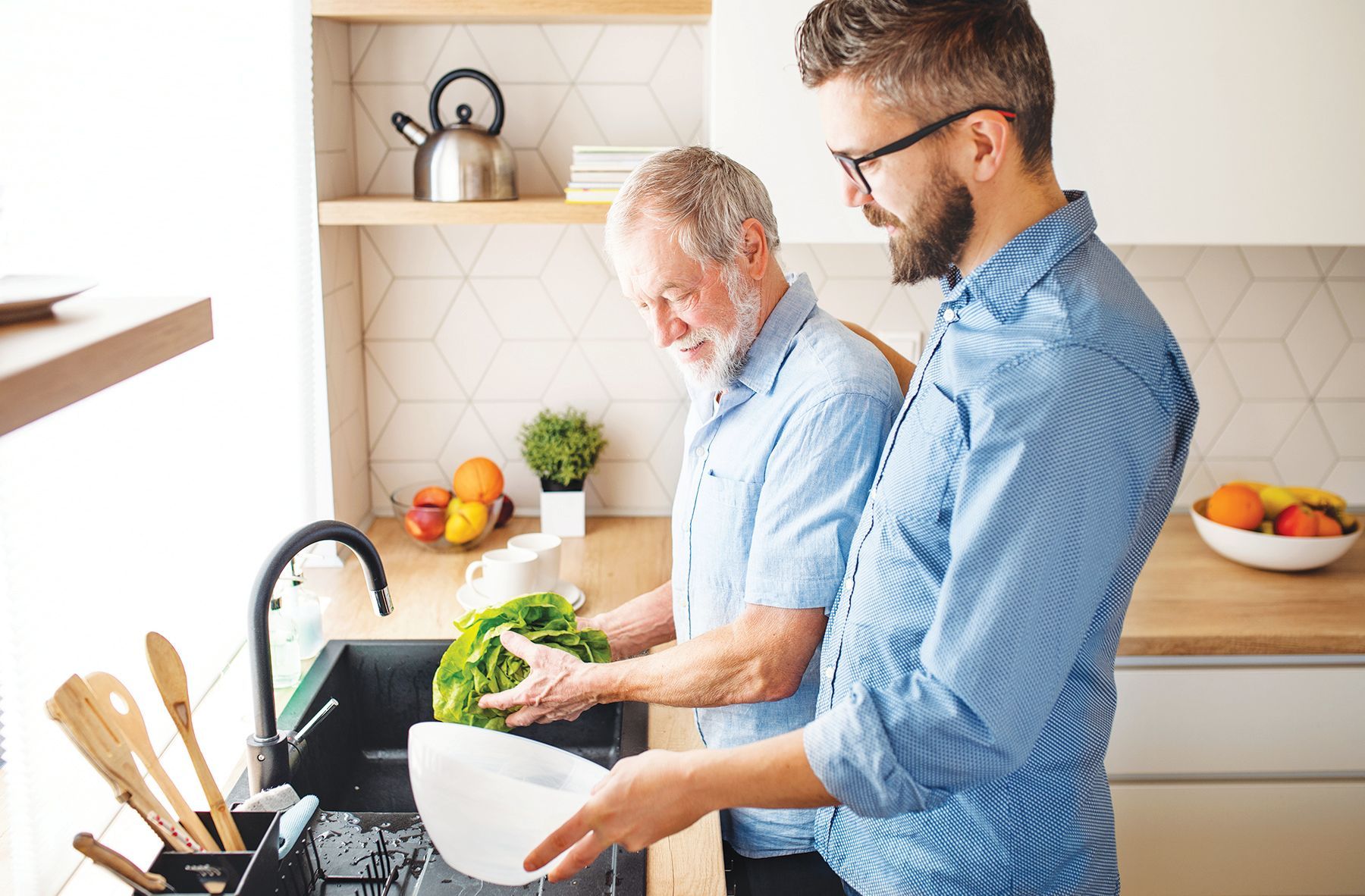Two people in a kitchen, one washing lettuce in a sink while the other holds a bowl nearby to help.