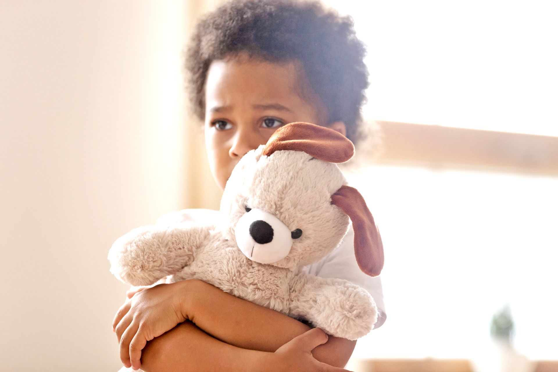A child holding a beige plush toy, looking thoughtfully toward the side in a brightly lit, soft-focus setting.