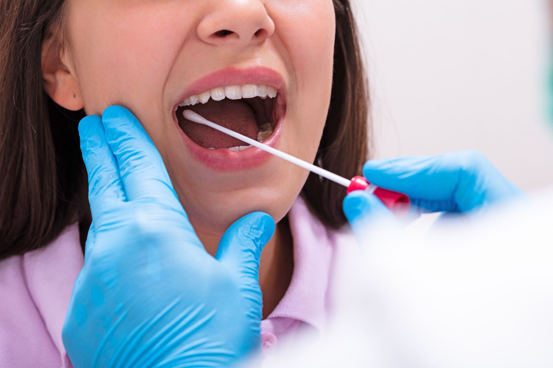 A healthcare worker wearing blue gloves uses a swab to collect a sample from a person’s throat.
