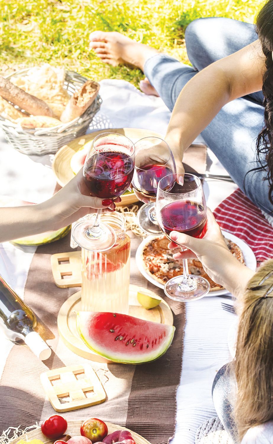 People clink glasses of red wine at a sunny outdoor picnic featuring a basket of bread, a watermelon slice, and fruit.
