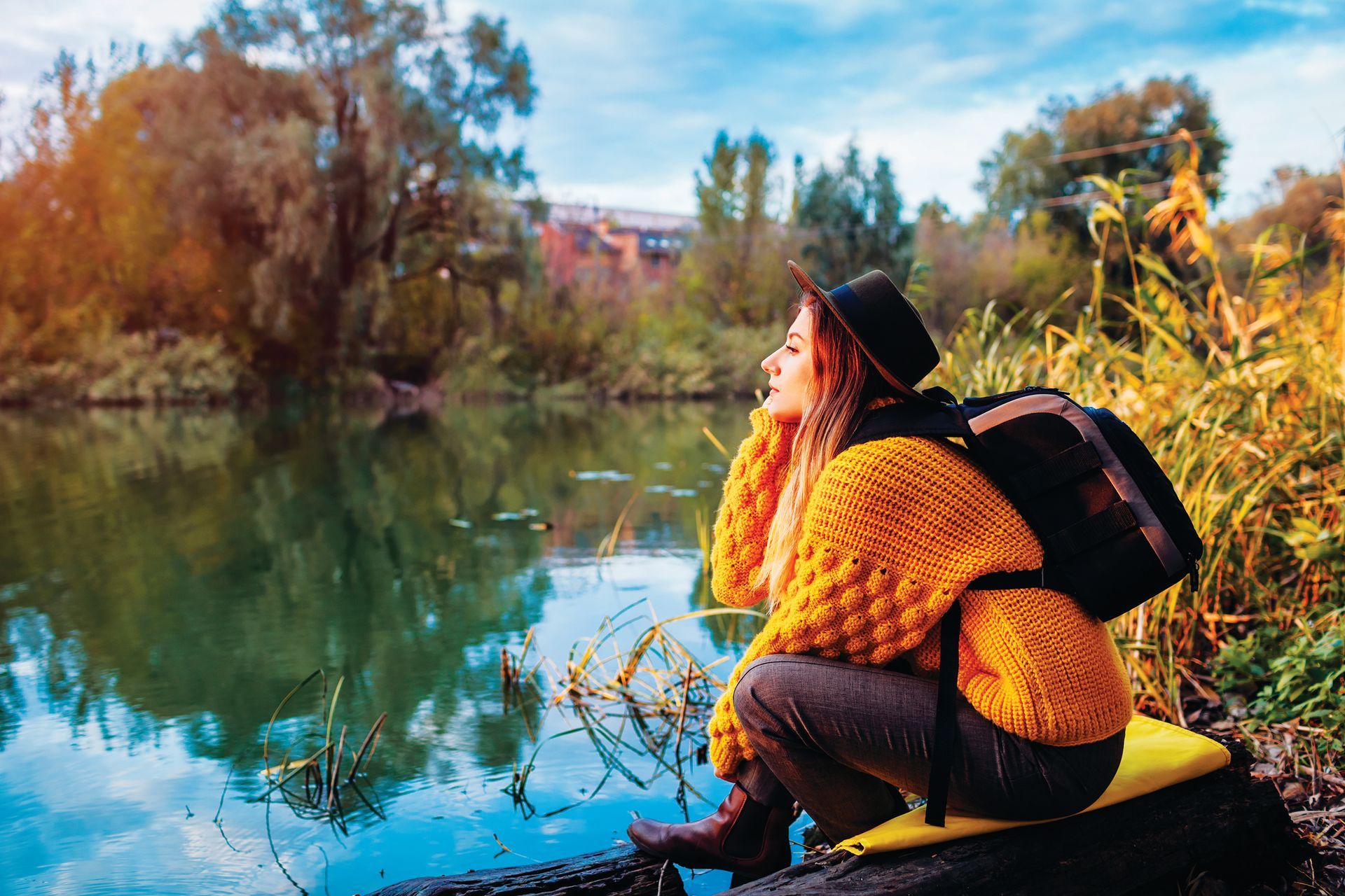 A person in a yellow sweater and hat sits by a reflective, calm river, surrounded by trees and tall grasses in autumn.