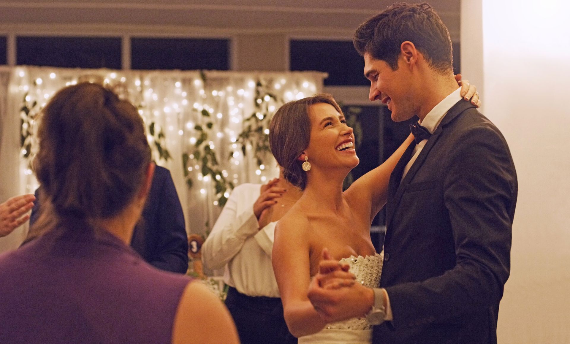 A bride and groom smile at each other while dancing at a reception with soft string lights in the background.