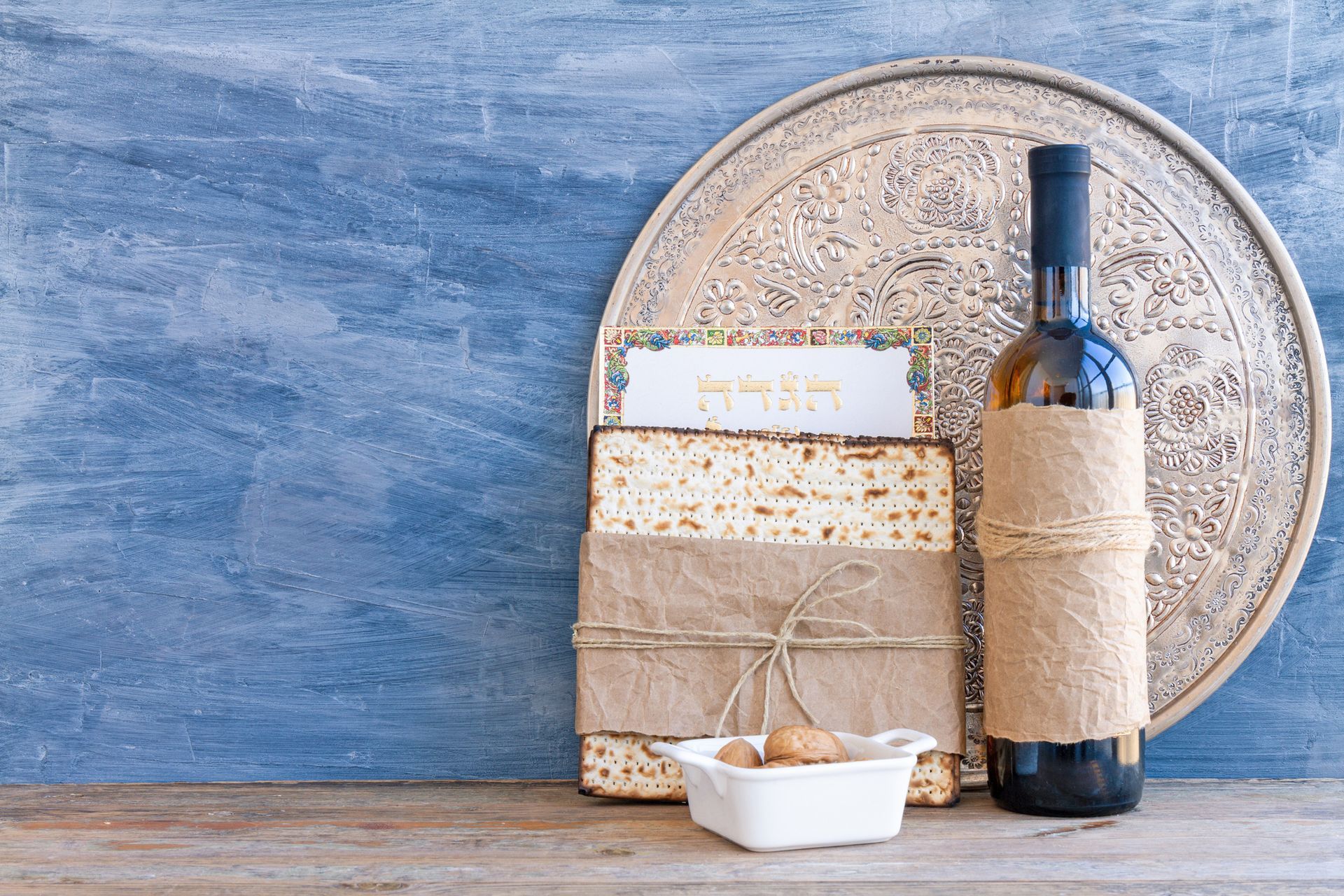 Matzah, a bottle of wine, and a small dish of nuts arranged on a wooden surface against a rustic blue background.