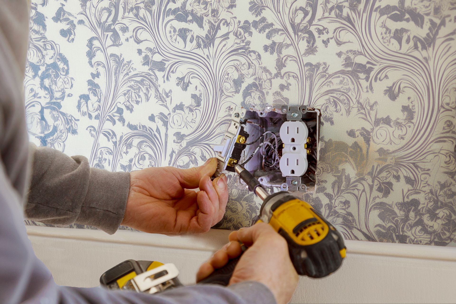 A person uses a power drill to install an electrical outlet into a wall covered in gray and white patterned wallpaper.