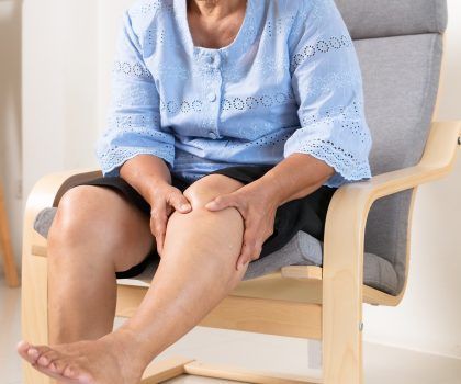A person sitting in a chair, holding their knee with both hands as if in pain.