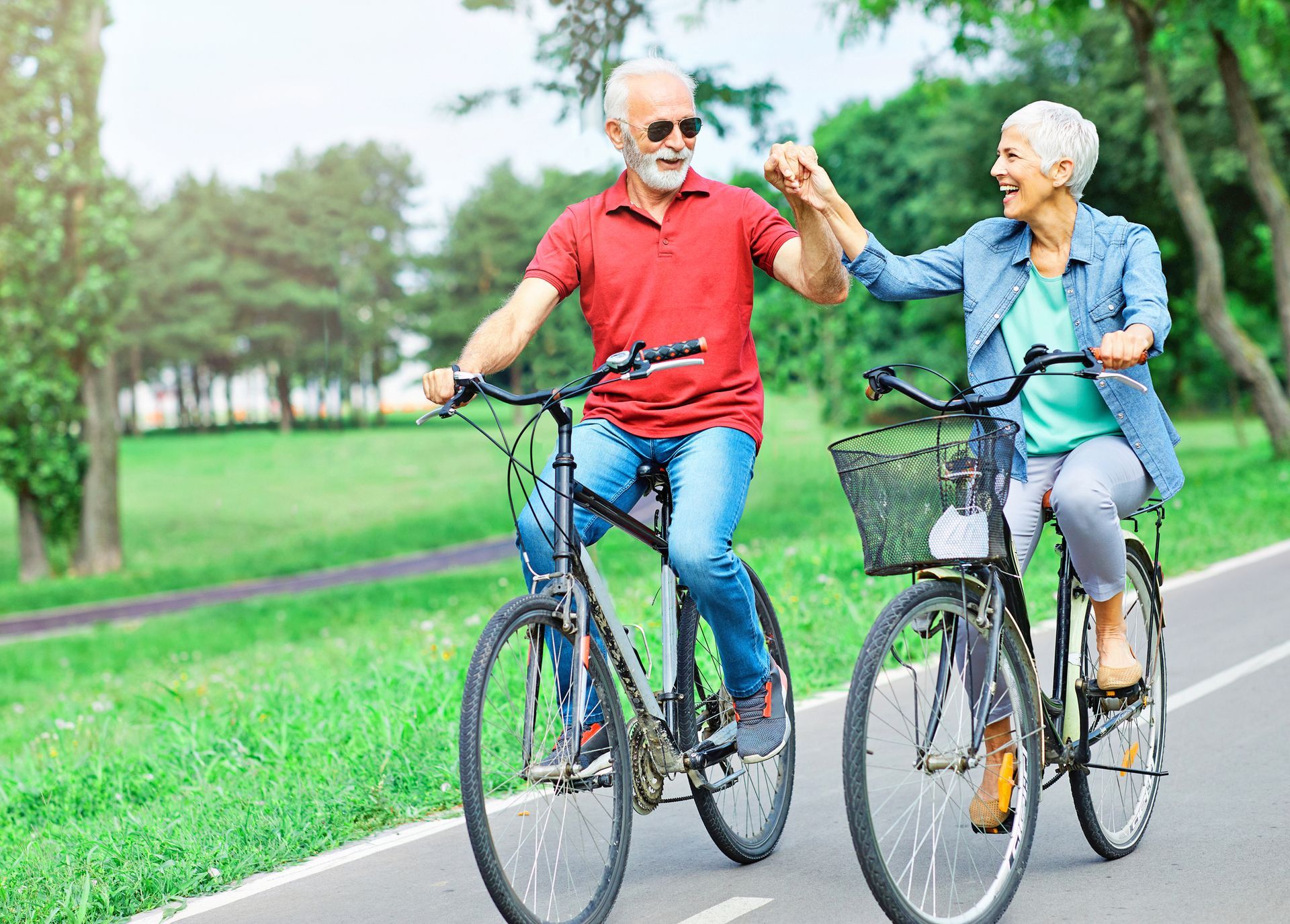 An active couple rides bicycles along a paved park path, smiling and holding hands while cycling side-by-side.
