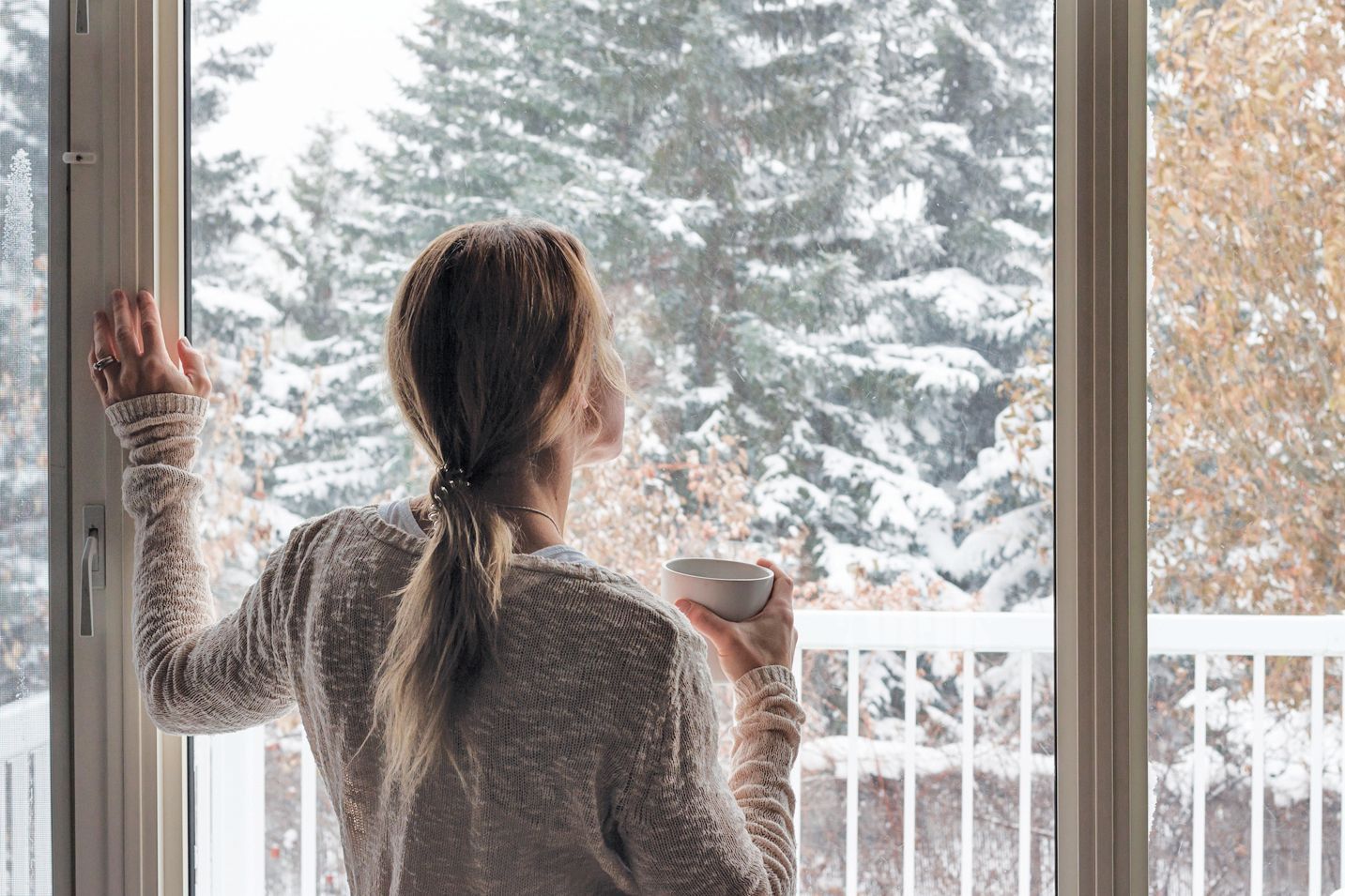 A person in a sweater stands at a window, holding a mug and looking out at a snow-covered forest.