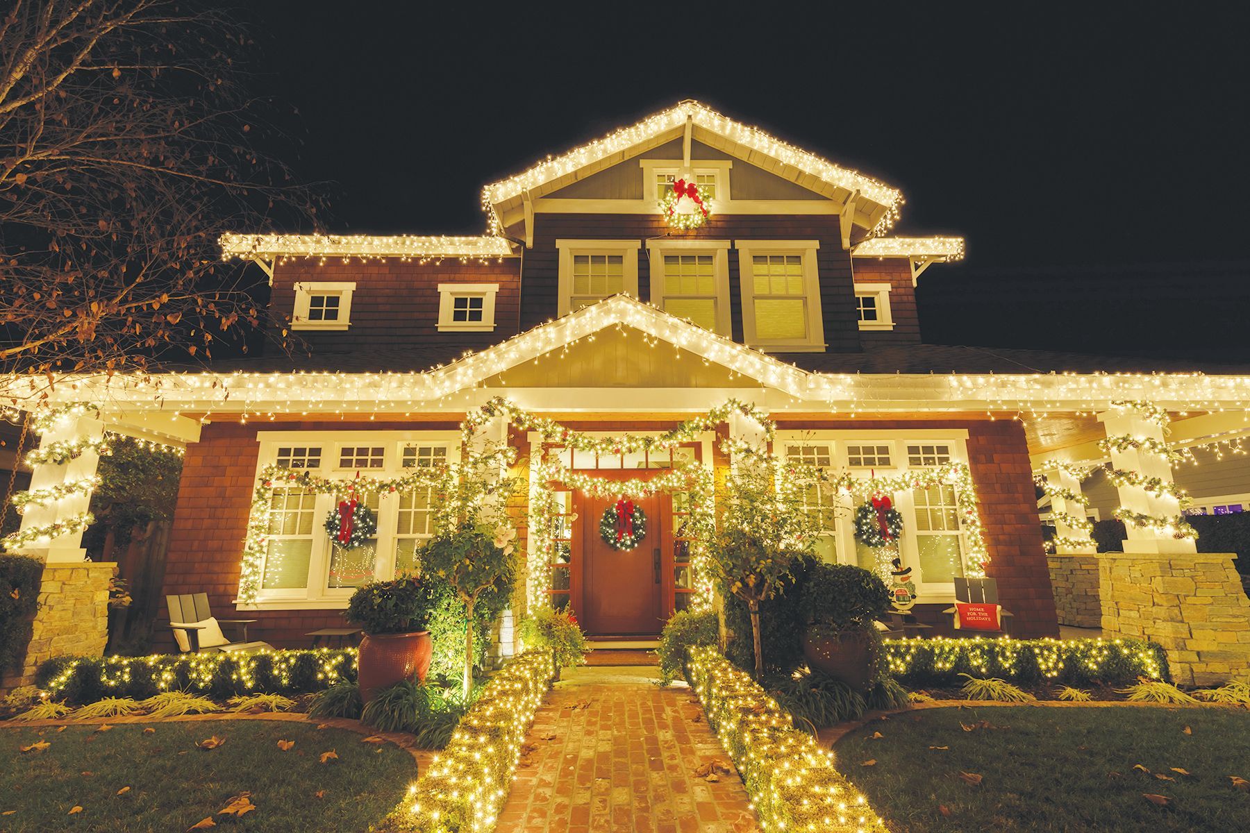 A two-story house at night, decorated with glowing warm white Christmas lights outlining its roof, porch, and walkway.