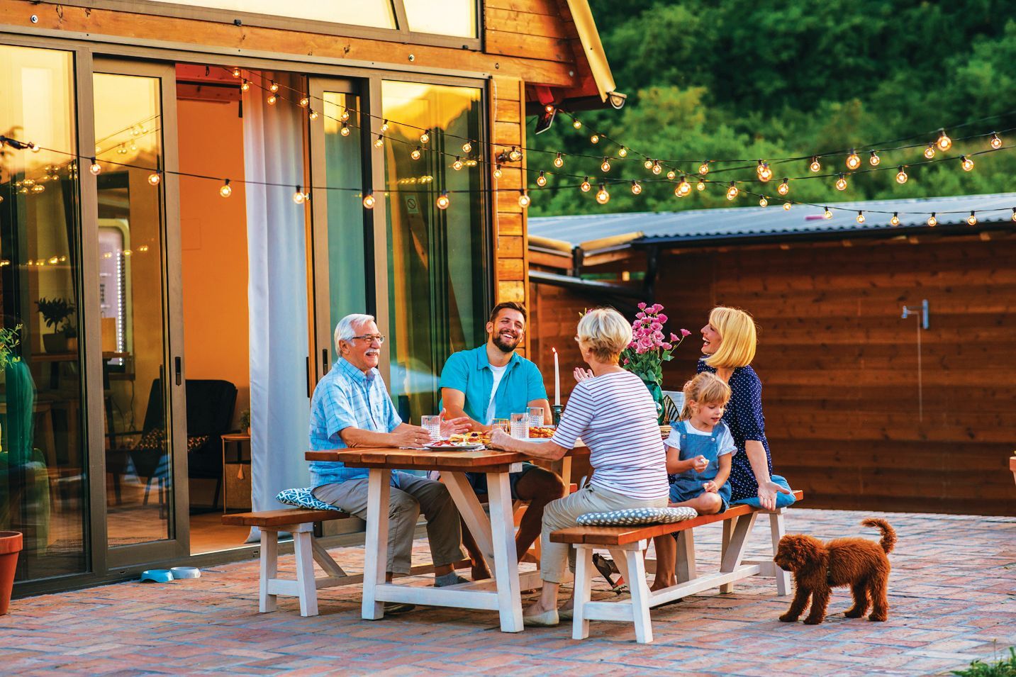 A group sits at an outdoor table on a stone patio by a wooden cabin, enjoying a meal under string lights with a small dog.