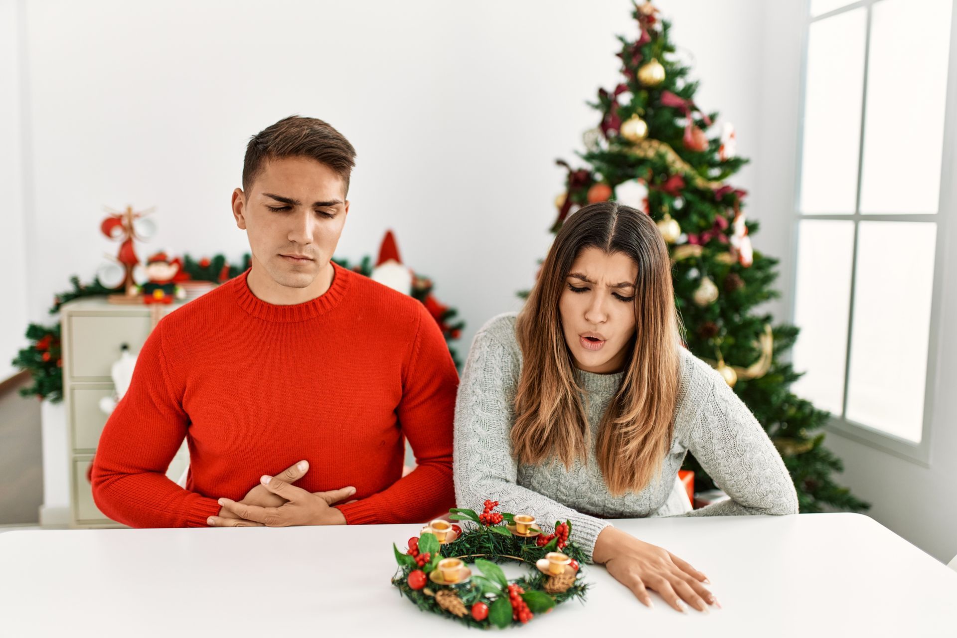 A man in a red sweater and a woman in a grey sweater look unhappy while sitting at a table with a small Christmas wreath.