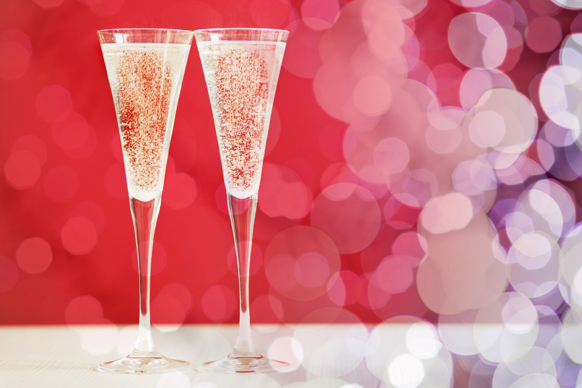 Two champagne flutes filled with sparkling wine against a festive red background with soft, blurry white bokeh lights.
