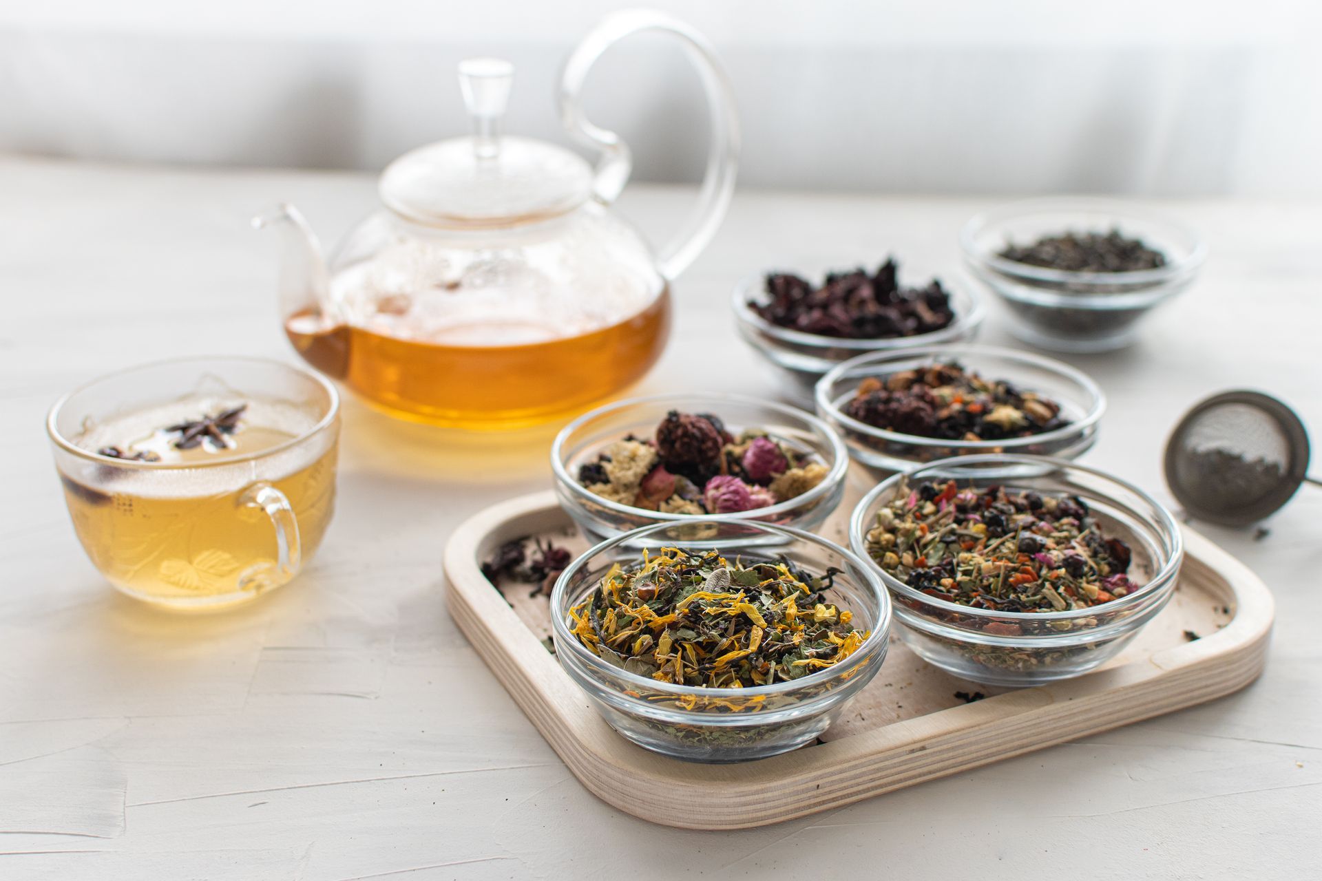 A glass teapot and cup filled with golden tea, beside small glass bowls of various loose-leaf teas on a wooden tray.