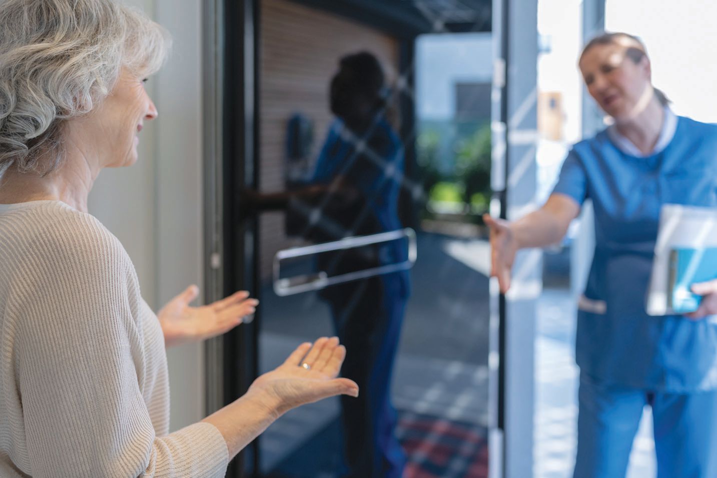A healthcare worker in blue scrubs opens a glass door for an individual, gesturing for them to enter.