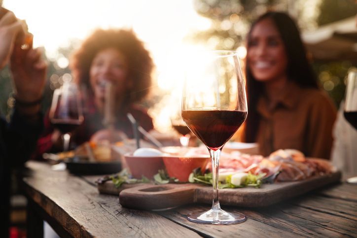 People sharing a meal and wine on a wooden table at sunset, with a glass of red wine in the foreground.