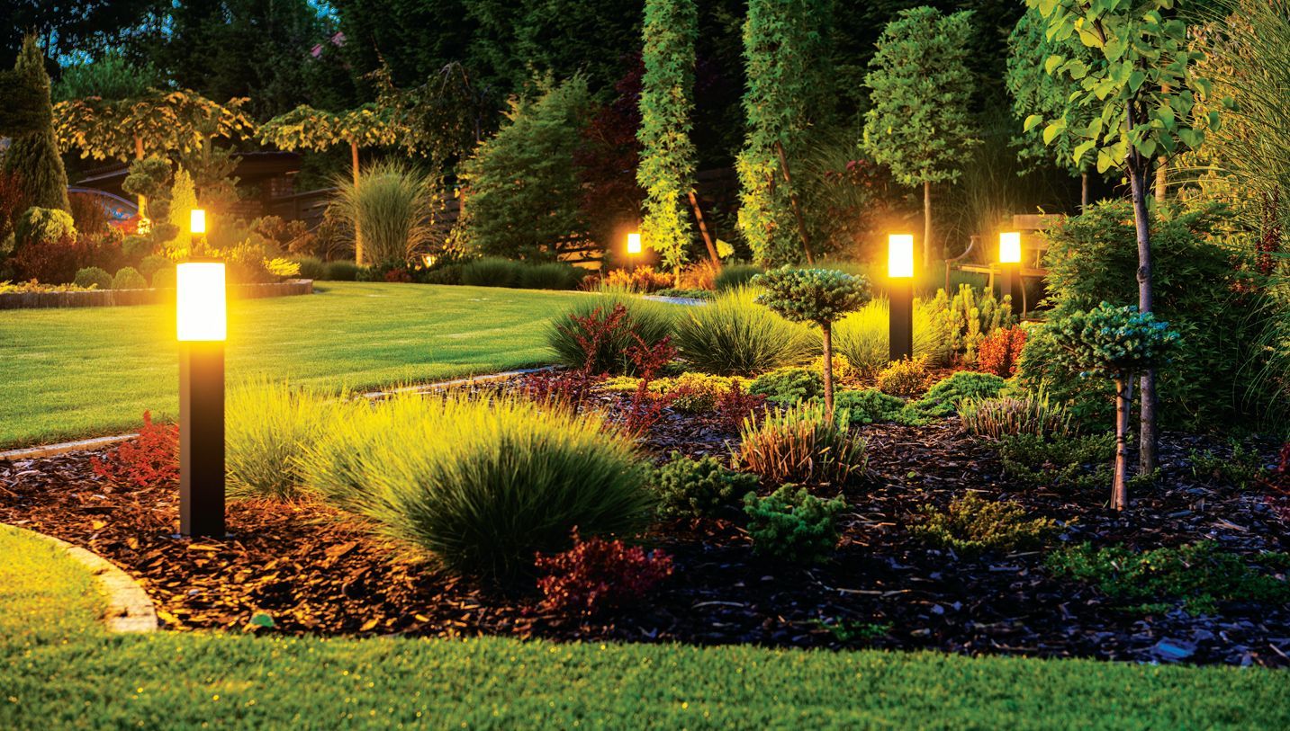 Illuminated garden path at dusk featuring several pillar-style lights shining on bushes, mulch, and trees.