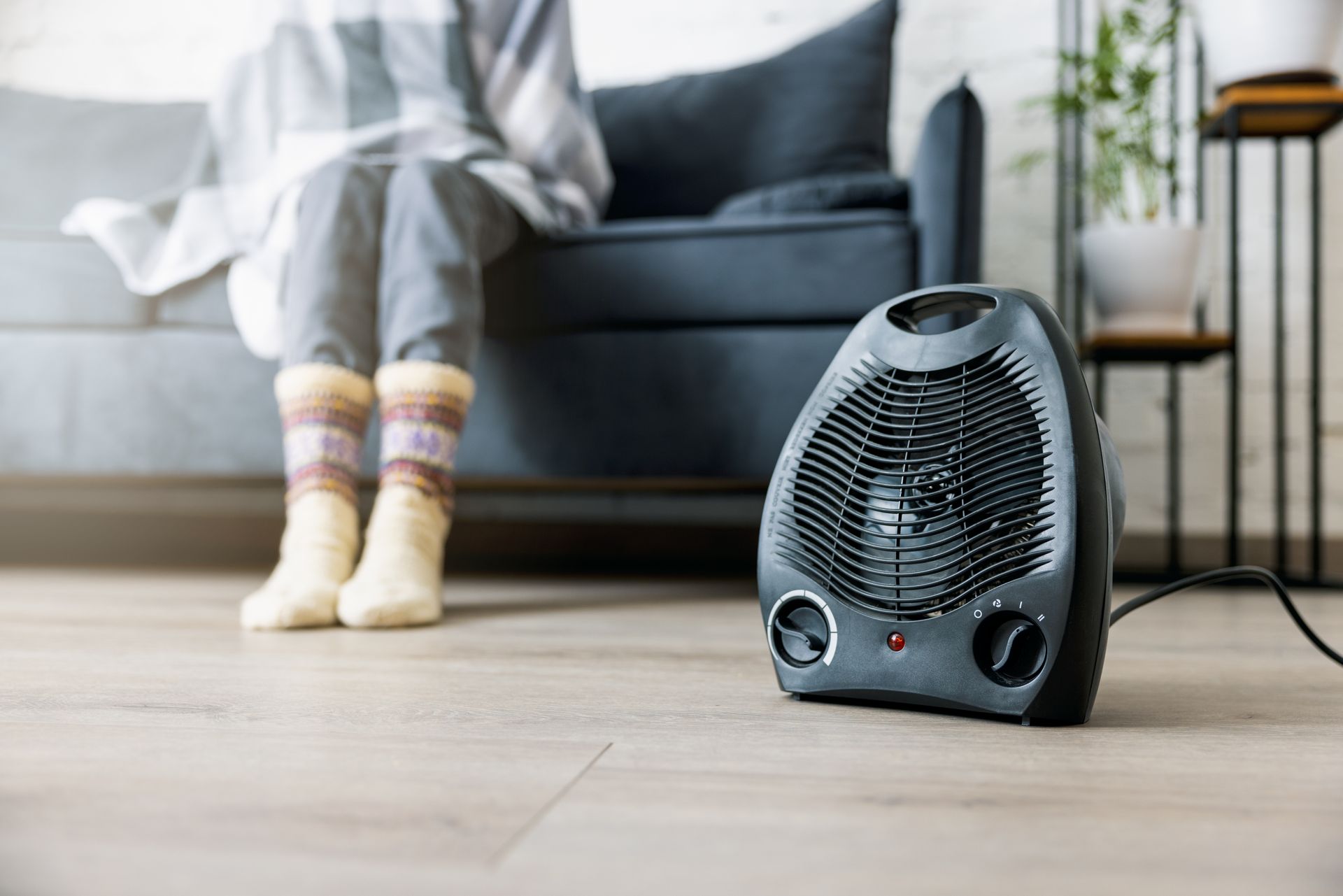 A small electric space heater sits on a wood floor in front of someone sitting on a sofa wrapped in a blanket.