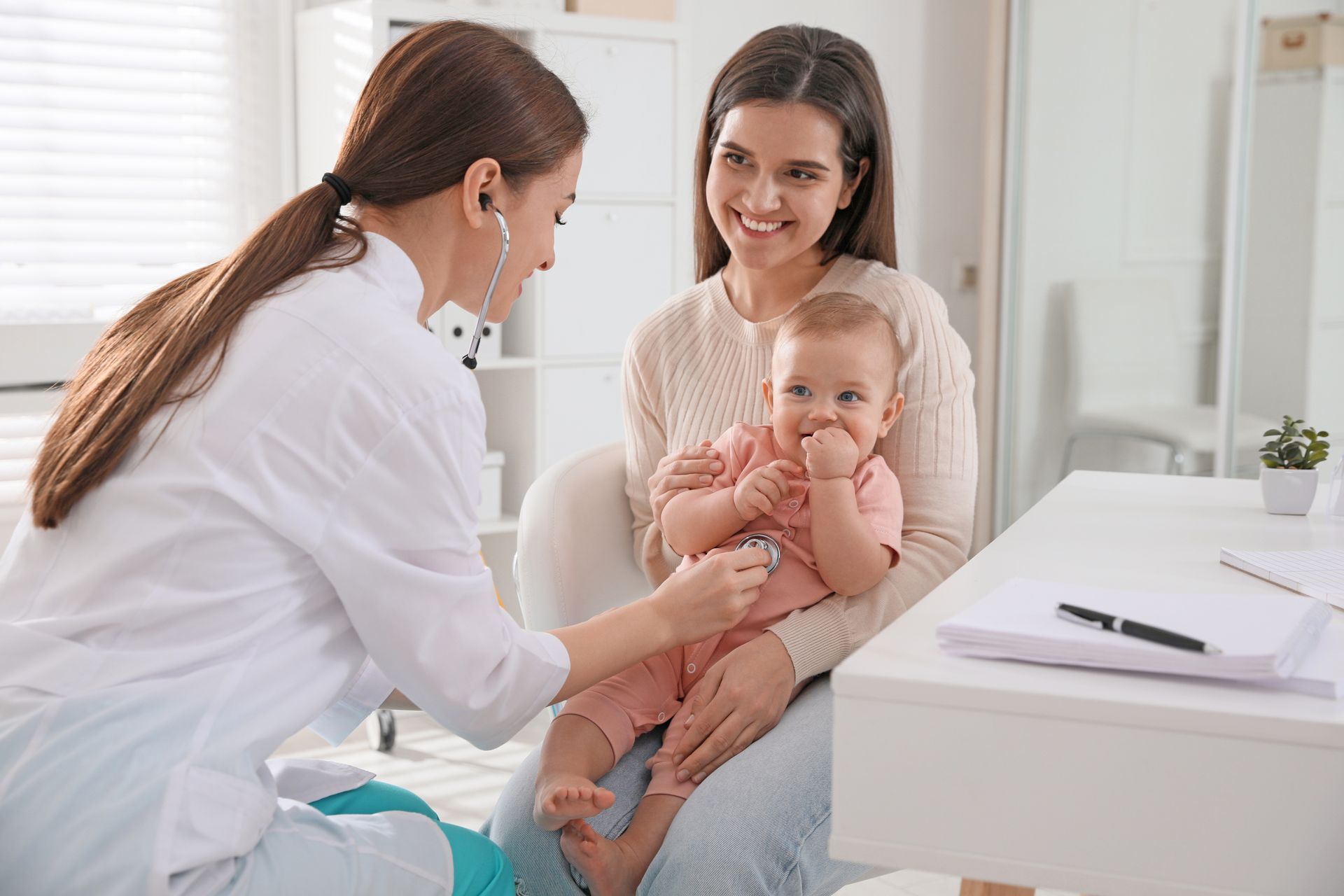 A healthcare professional uses a stethoscope to examine a young child sitting on a parent's lap in a clinic.