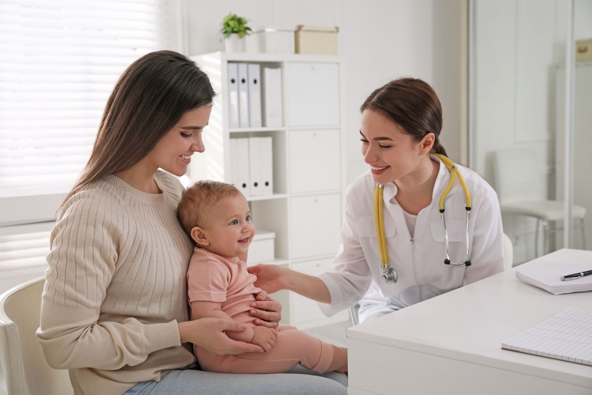 A pediatrician with a stethoscope smiles at a baby sitting on a parent's lap in a bright, modern medical office.
