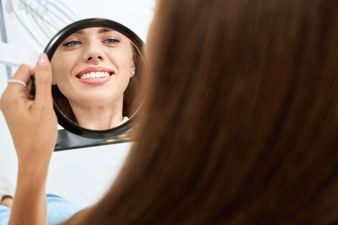 A person holds a small, round mirror, looking at their reflection and smiling to show their teeth.